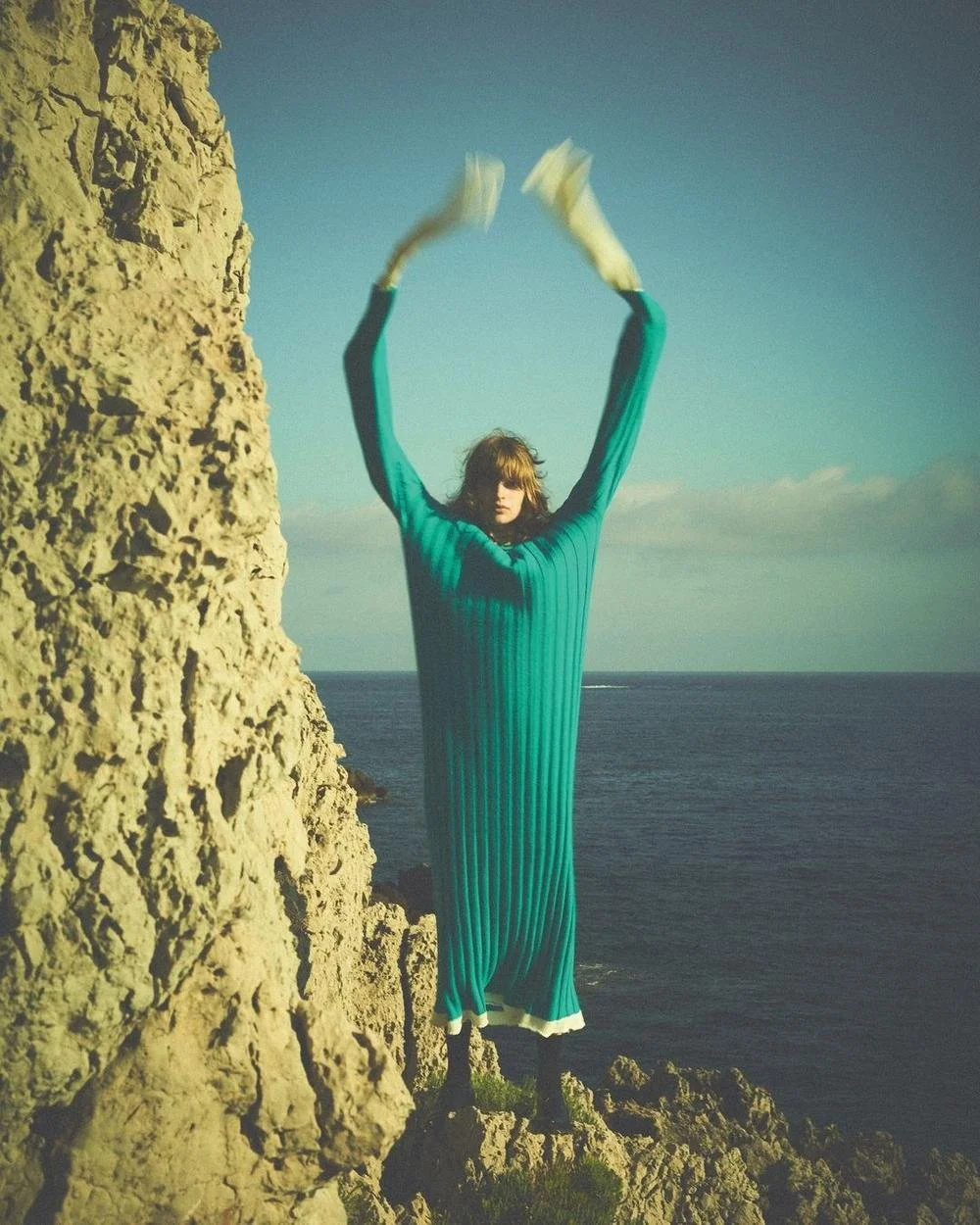 Person with long hair in a teal dress standing on rocky cliff with arms raised and blurred motion of hands, overlooking ocean with clear sky.