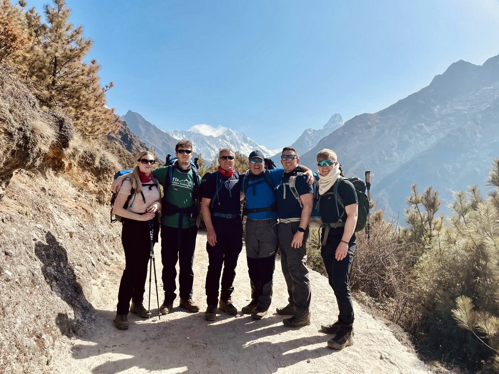 Group of six hikers in mountains with snow-capped peaks, wearing outdoor gear and sunglasses, standing on a dirt trail surrounded by pine trees and rocky terrain.