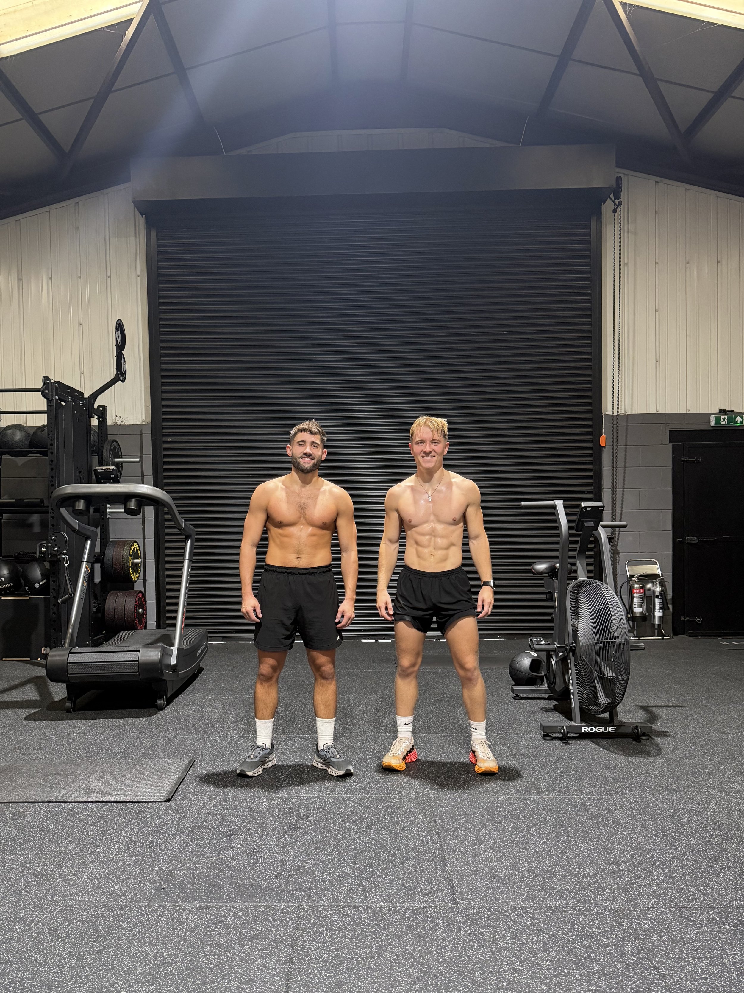 Two shirtless men standing in a gym, smiling at the camera. The gym has various workout equipment, a large black roll-up door, and a fan on the floor.