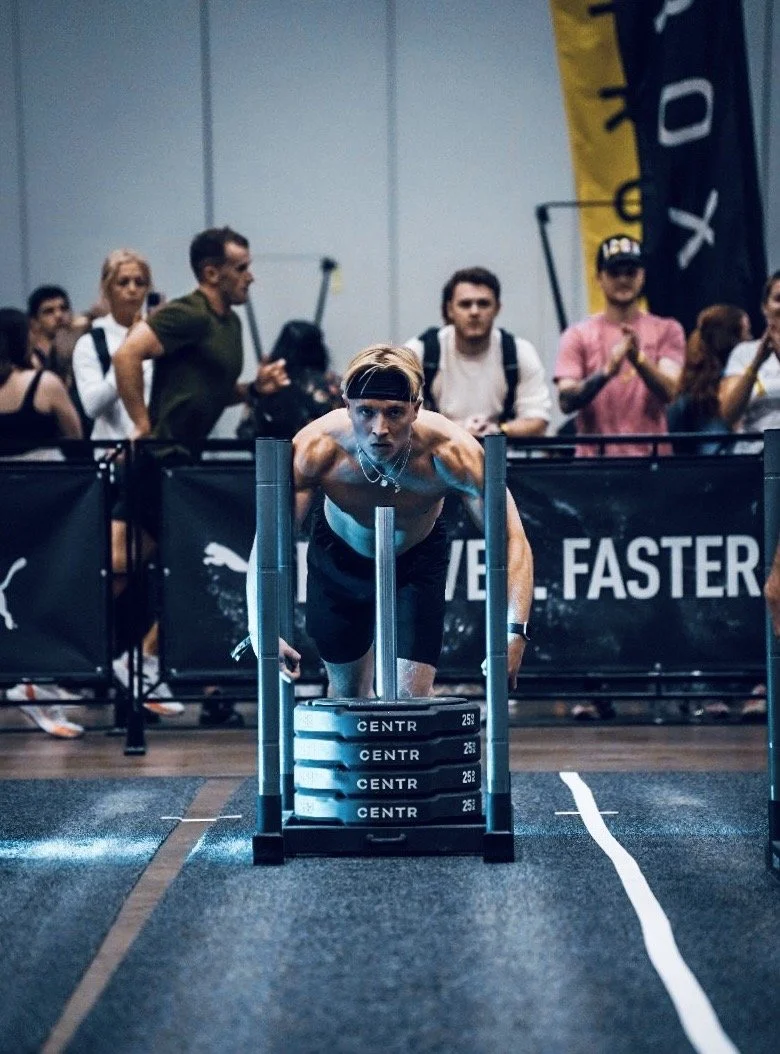 A man pushing a weighted sled at a fitness competition, with onlookers surrounding him.