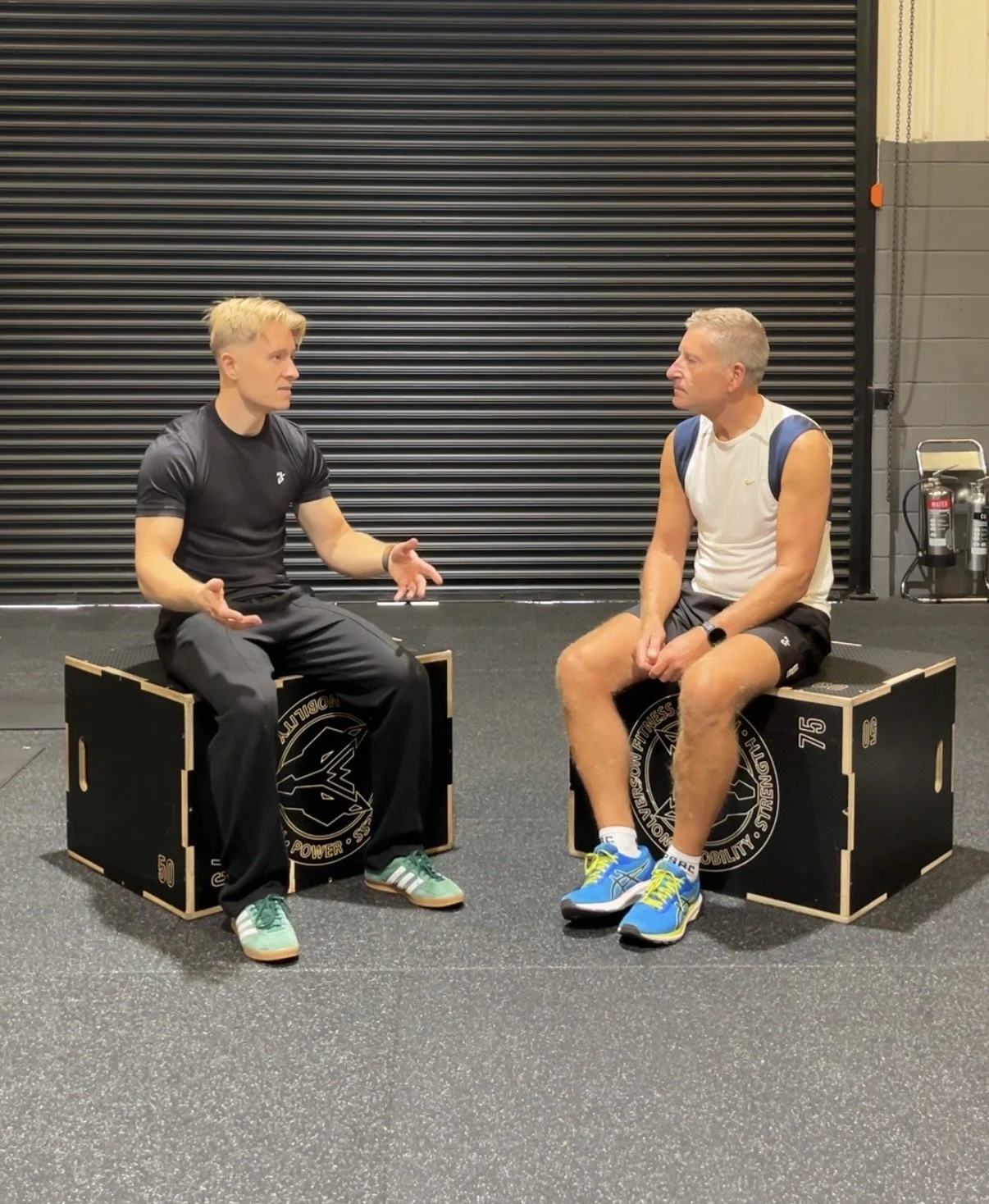 Two men sitting on workout boxes having a conversation at the gym.