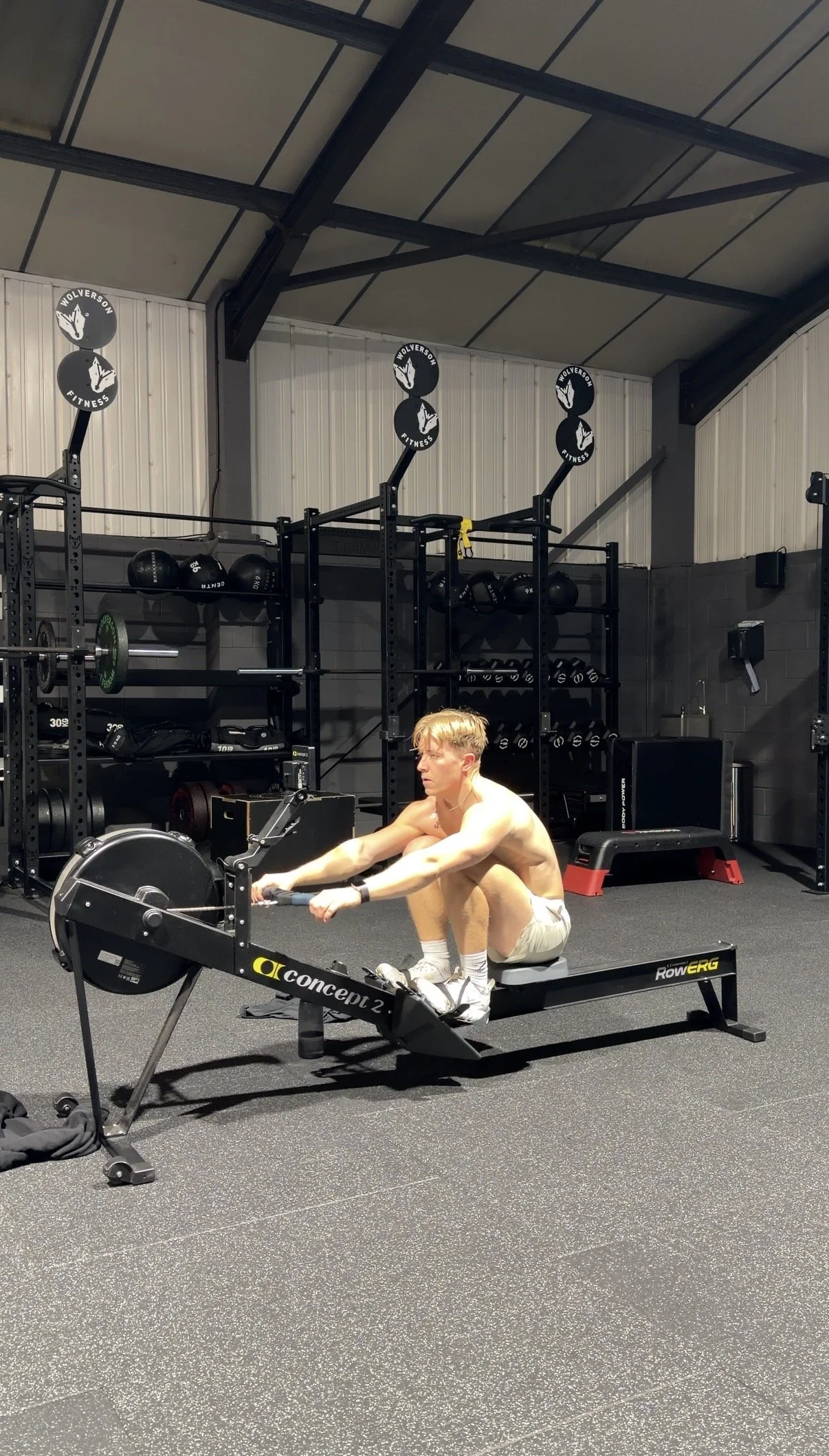 A shirtless young man with blonde hair and white shorts sitting on a rowing machine in a gym, with weight racks and gym equipment in the background.