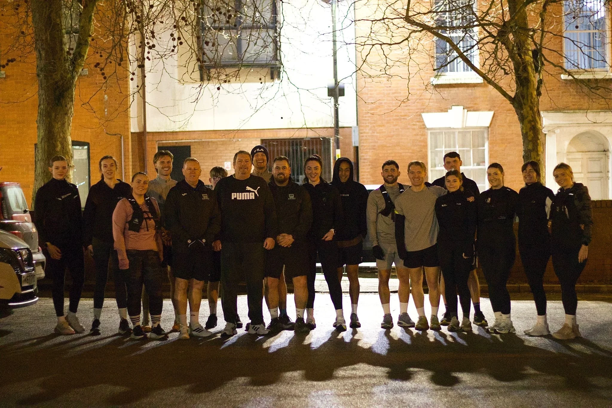 A group of people, mostly young adults, standing together outdoors at night in front of residential buildings, smiling for the camera. Two trees are visible in the street with bare branches, indicating a colder season. The group appears to be friends or teammates.