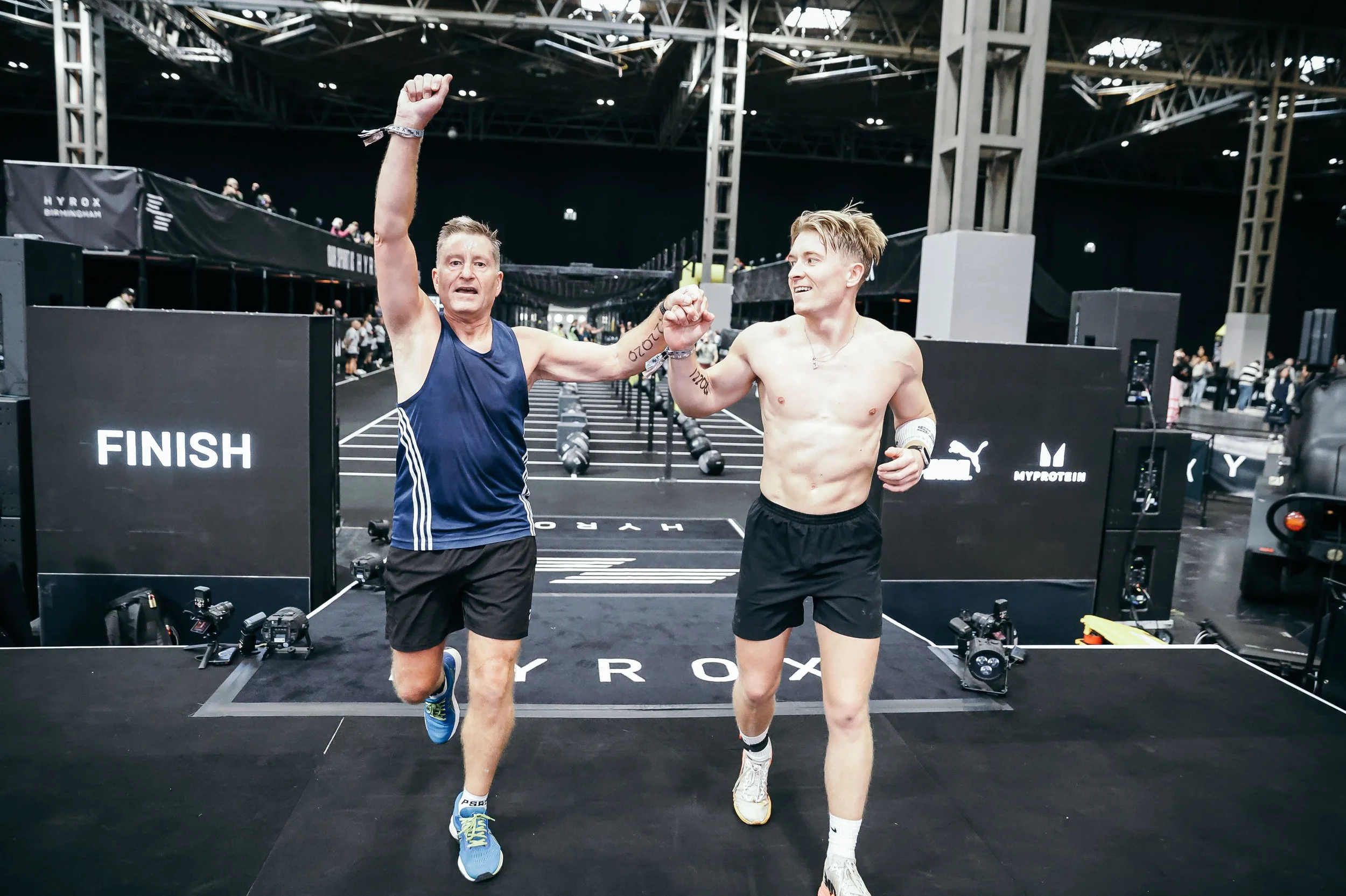 Two male athletes celebrating after a race at the finish line in an indoor sports arena. They are holding hands and raising their fists in victory. The gentleman holding his hand up has completed a Hyrox with Parkinsons.