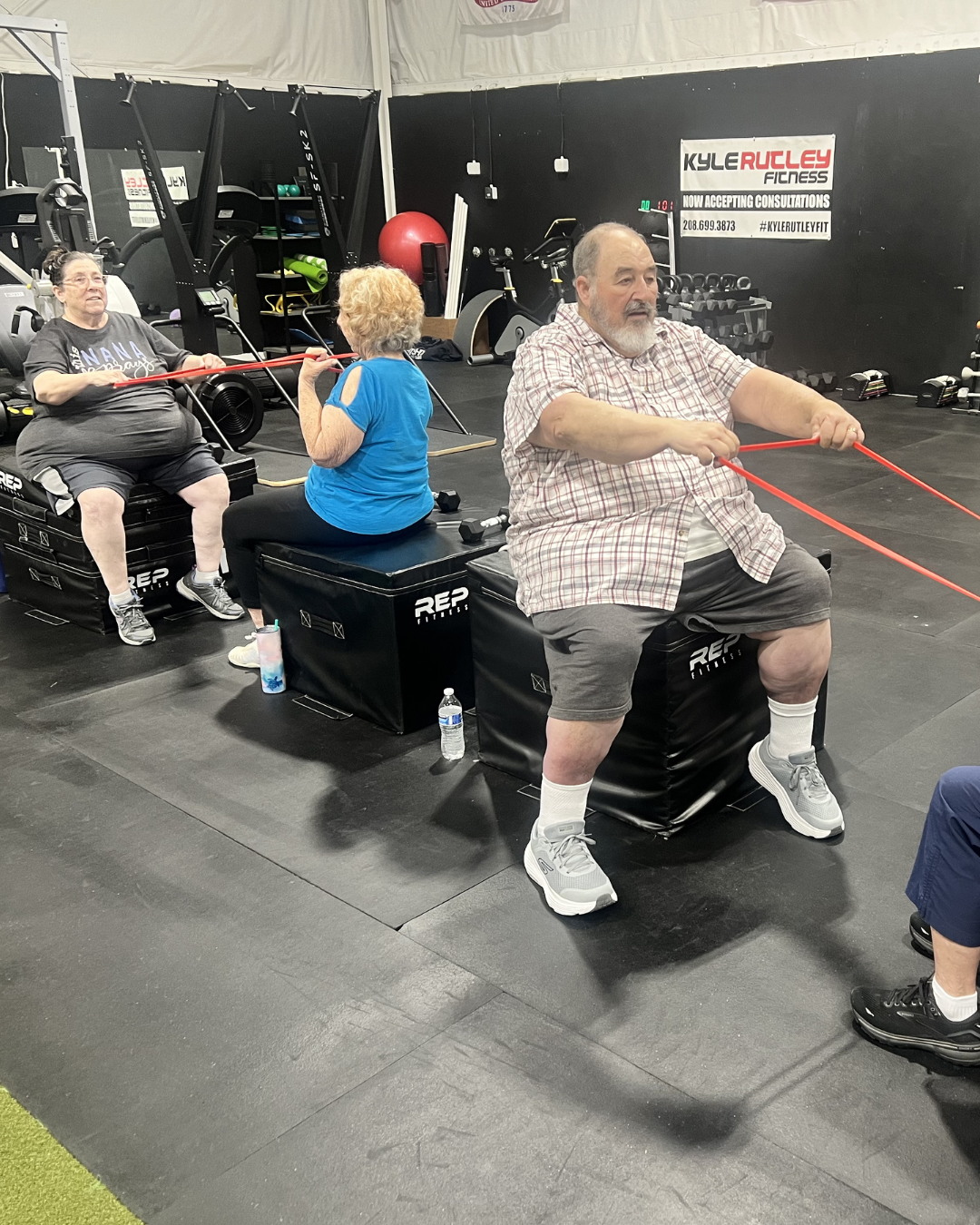 Three seniors working out in a gym, each sitting on a black padded box, using resistance bands for exercise, with gym equipment visible in the background.