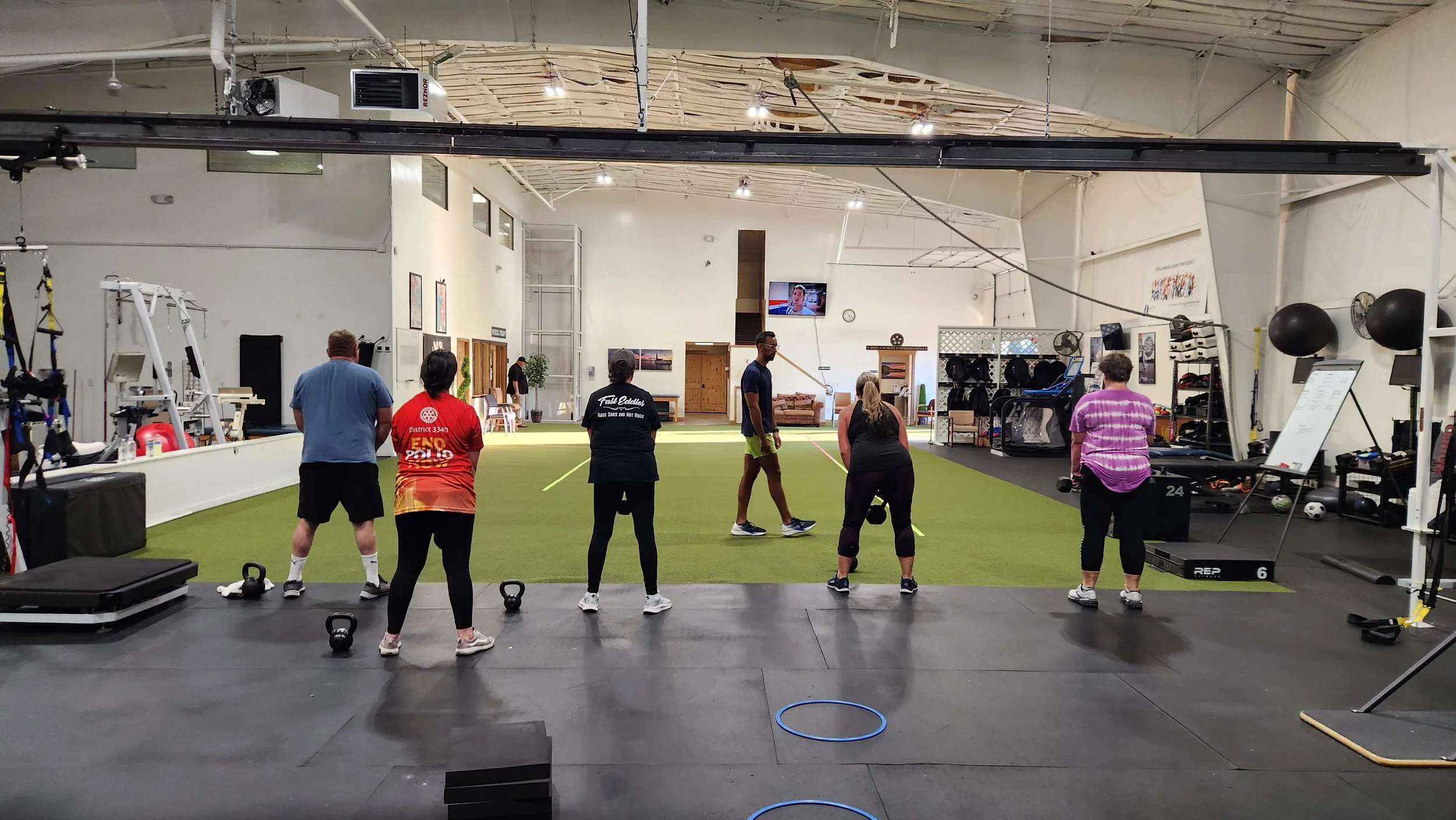 Group of people participating in a fitness class or workout session inside a gym, with exercise equipment and a large open workout area.