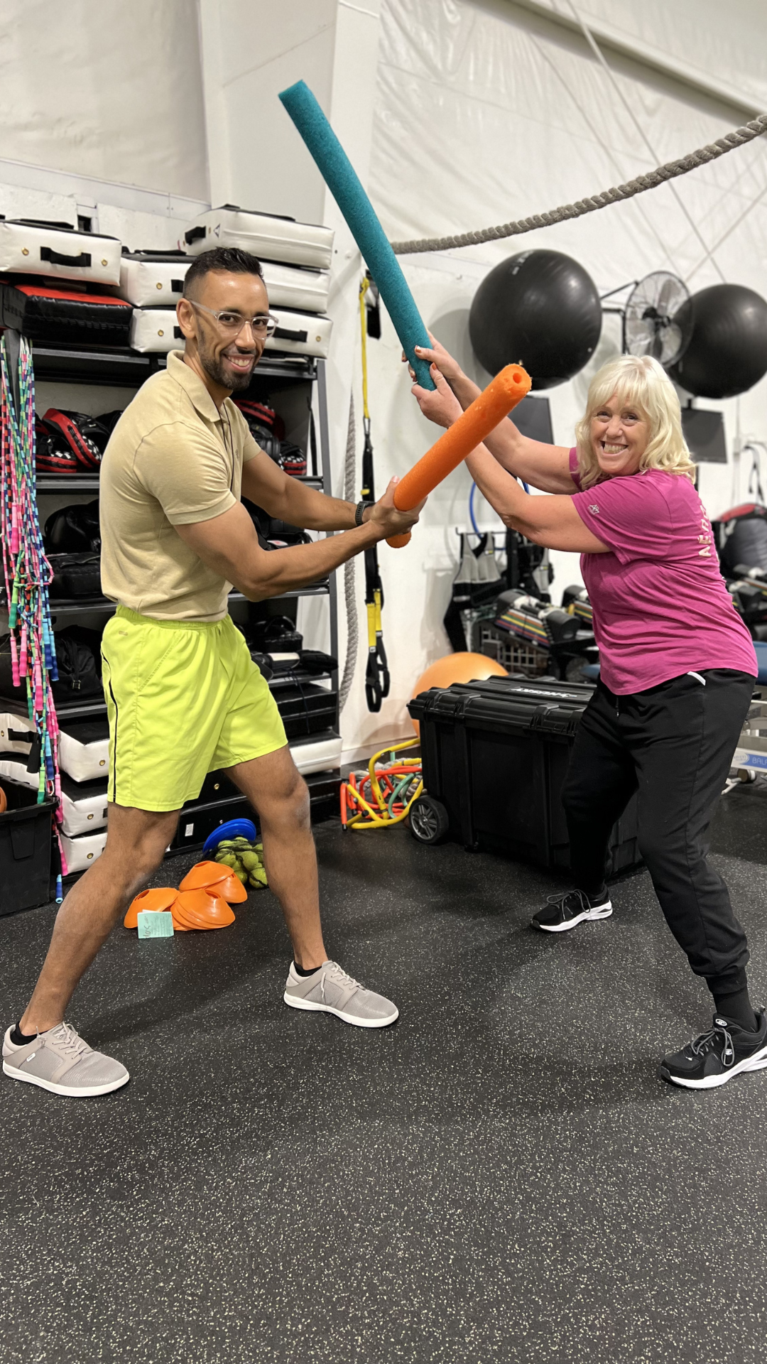 Two women are playfully dabbing foam sticks at each other in a gym. One woman is wearing yellow shorts and a beige shirt, and the other is wearing black pants and a pink shirt. Fitness equipment and supplies are visible in the background.