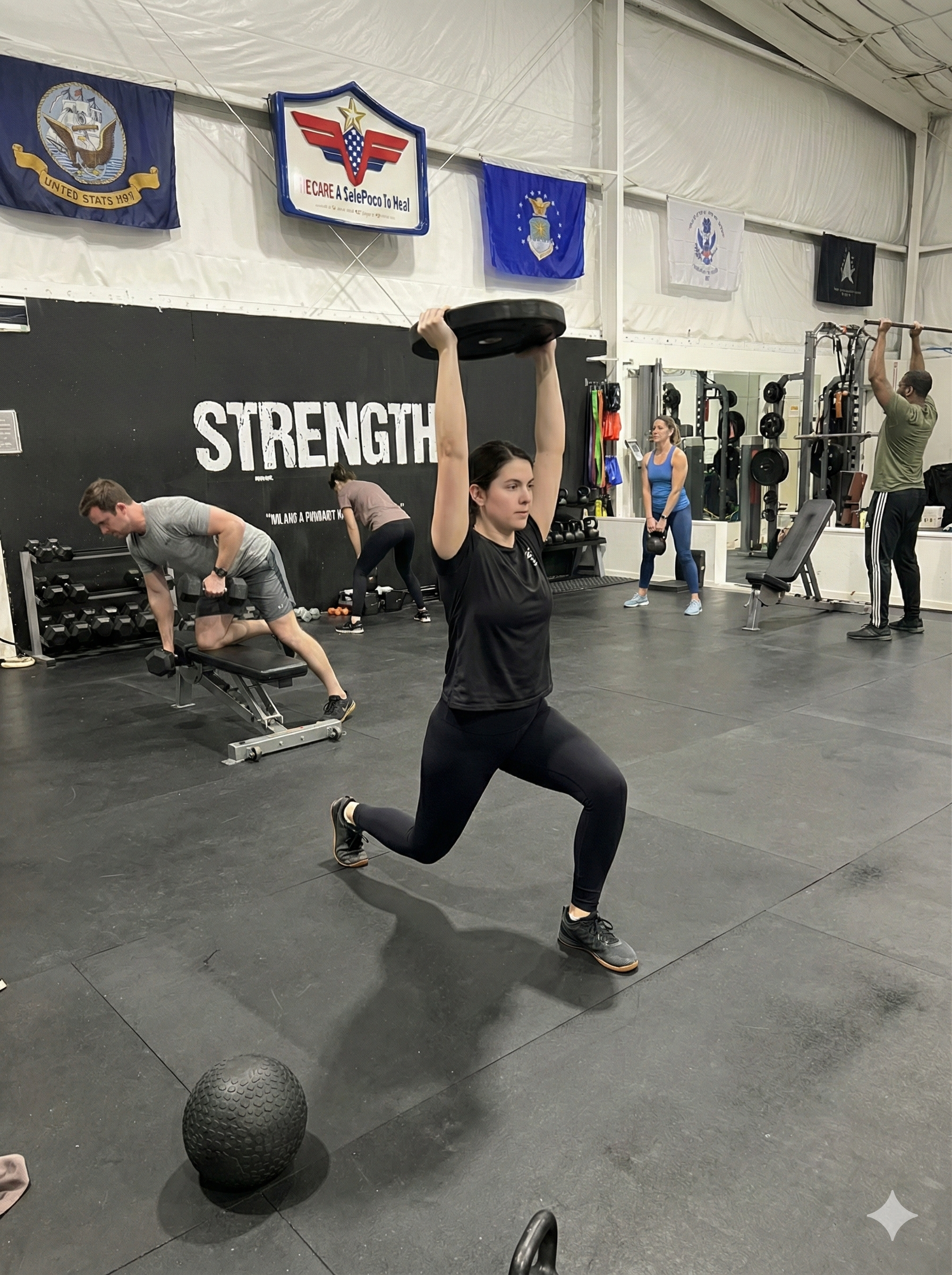 A woman in black workout clothing is performing a lunge while holding a weight overhead. She is in a gym with other people exercising, including a man bending over weights and a woman doing kettlebell swings, with various gym equipment and flags hanging on the wall.