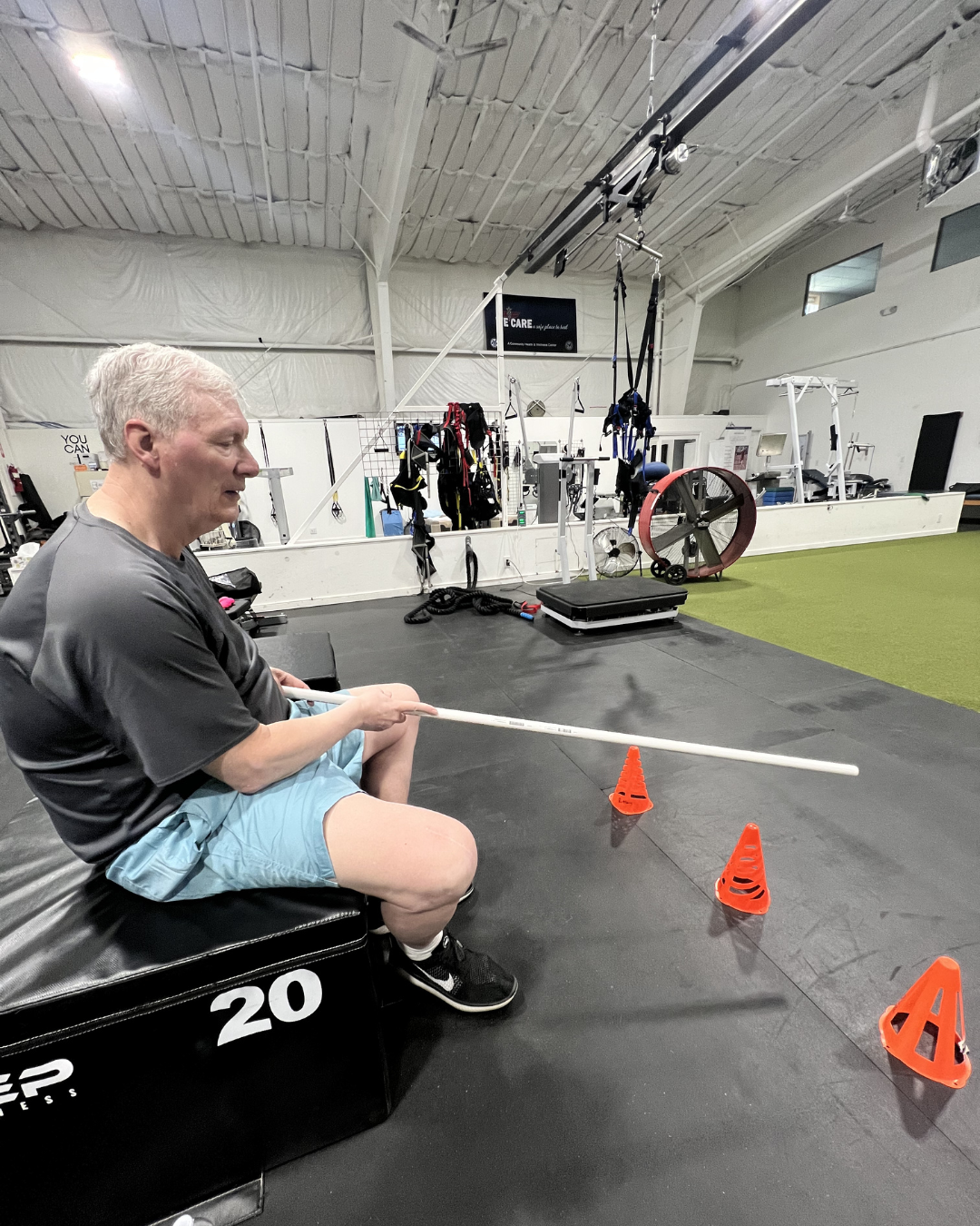 An older man sitting on a black workout box in a gym, holding a long white pvc pipe, with orange cones arranged in a line in front of him, in a gym with training equipment in the background.