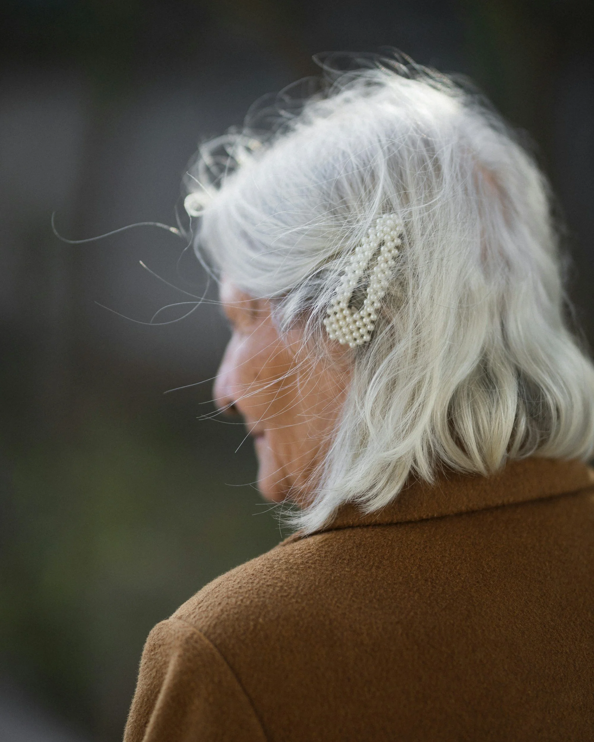 Close-up of a woman with long white hair wearing a pearl hair accessory and a brown coat, seen from the side, with a blurred background.