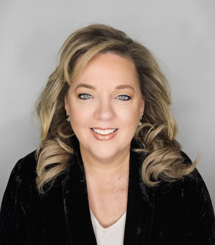 A smiling woman with blonde, wavy hair, wearing makeup, earrings, a white top, and a black velvet blazer, against a plain gray background.