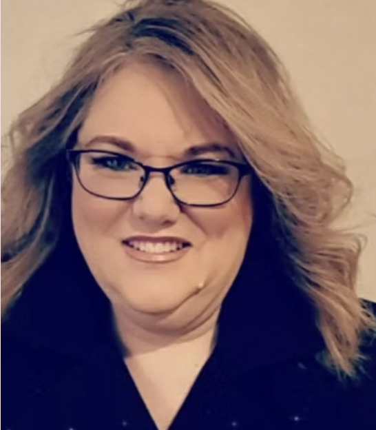 A woman with blonde, wavy hair wearing glasses and a dark top, smiling at the camera.