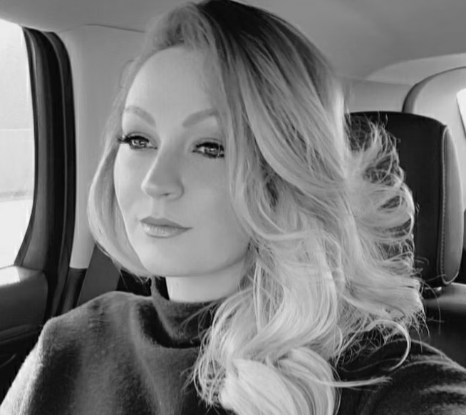 A woman with long, wavy blonde hair sitting in the backseat of a car, looking out the window.