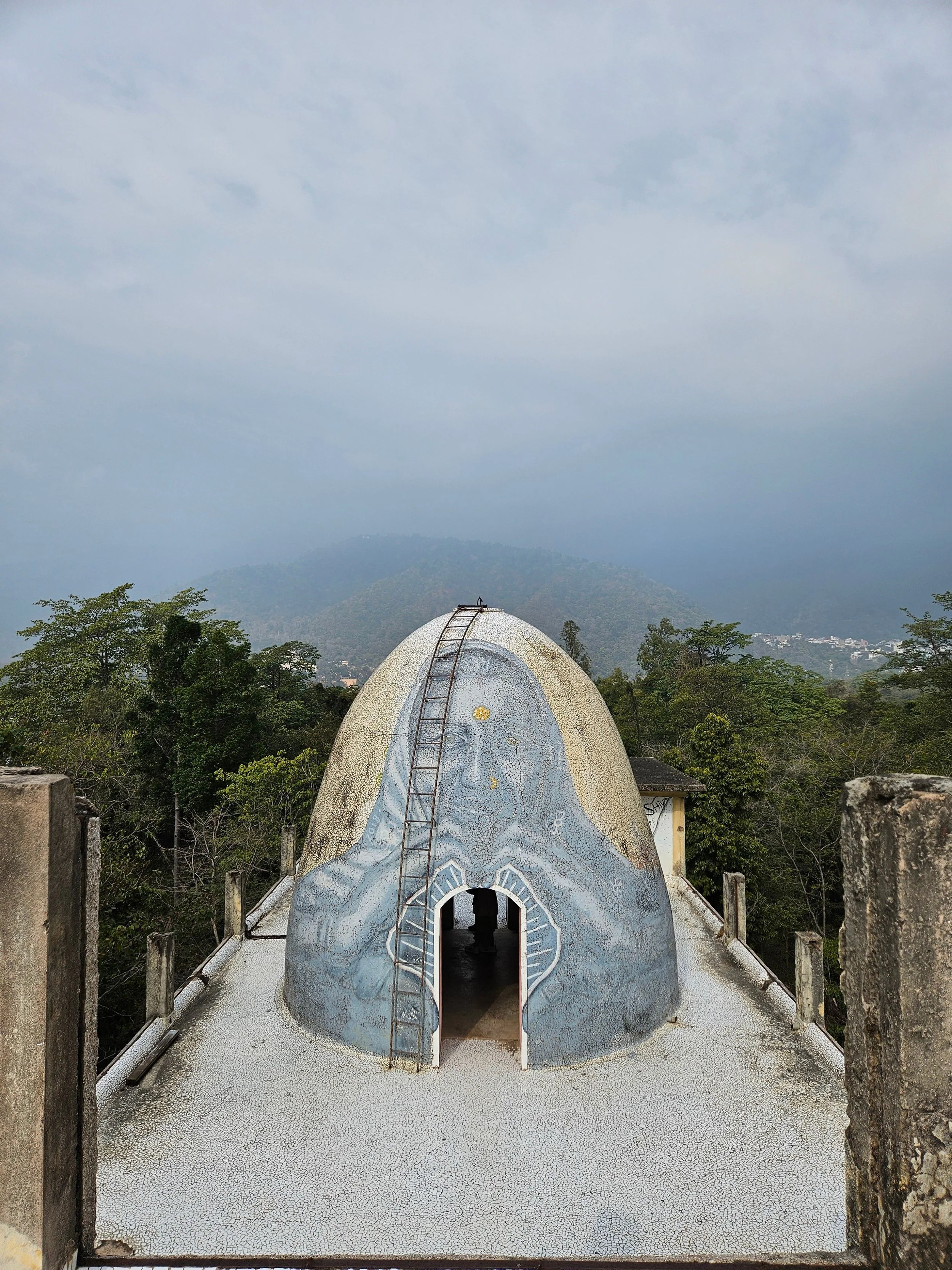A dome-shaped structure with an arched entrance and a ladder on top, surrounded by a railing, with trees and a mountain in the background under a cloudy sky.
