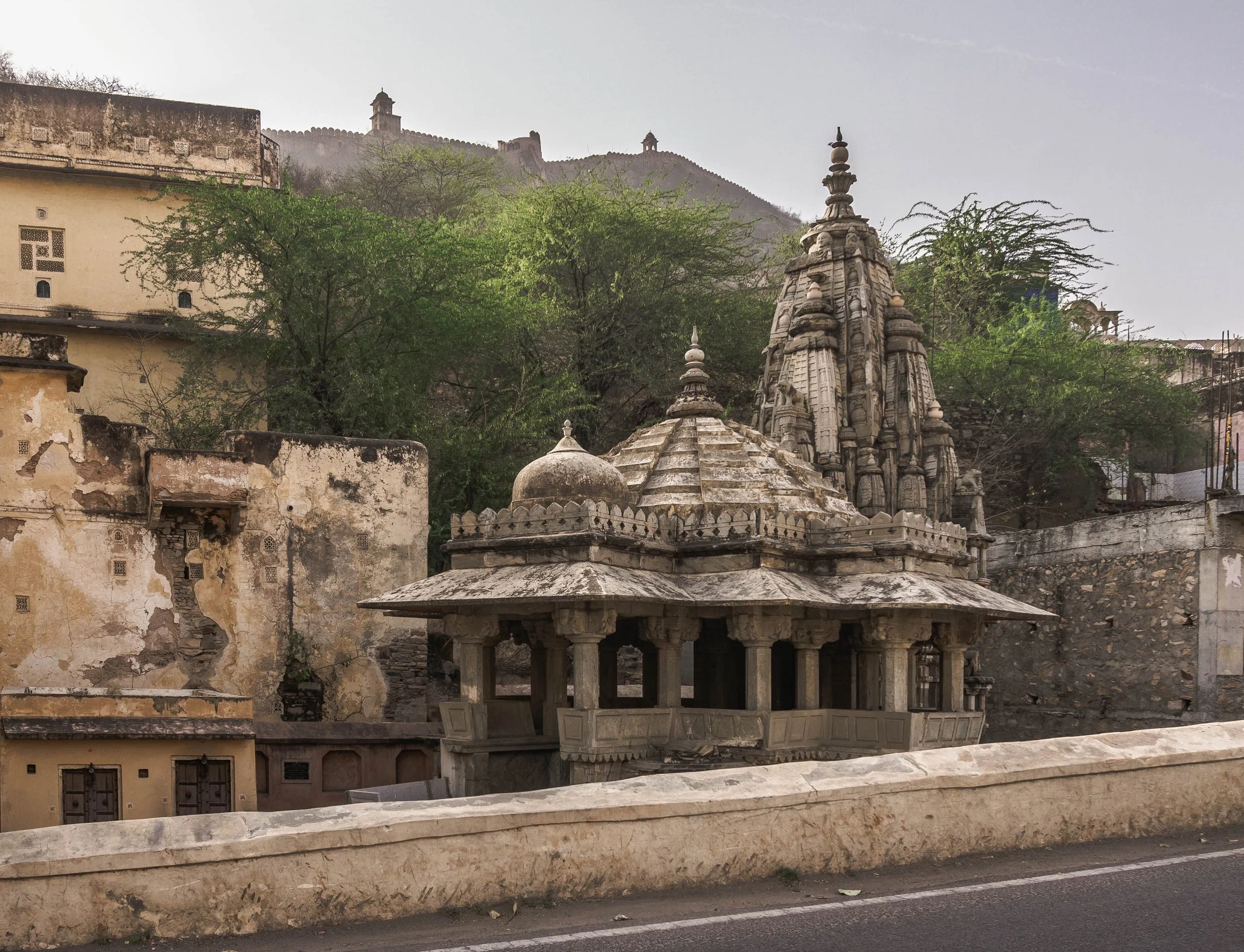 Ancient stone temple structure with detailed carvings, surrounded by trees, with a fort and hill in the background under an overcast sky.