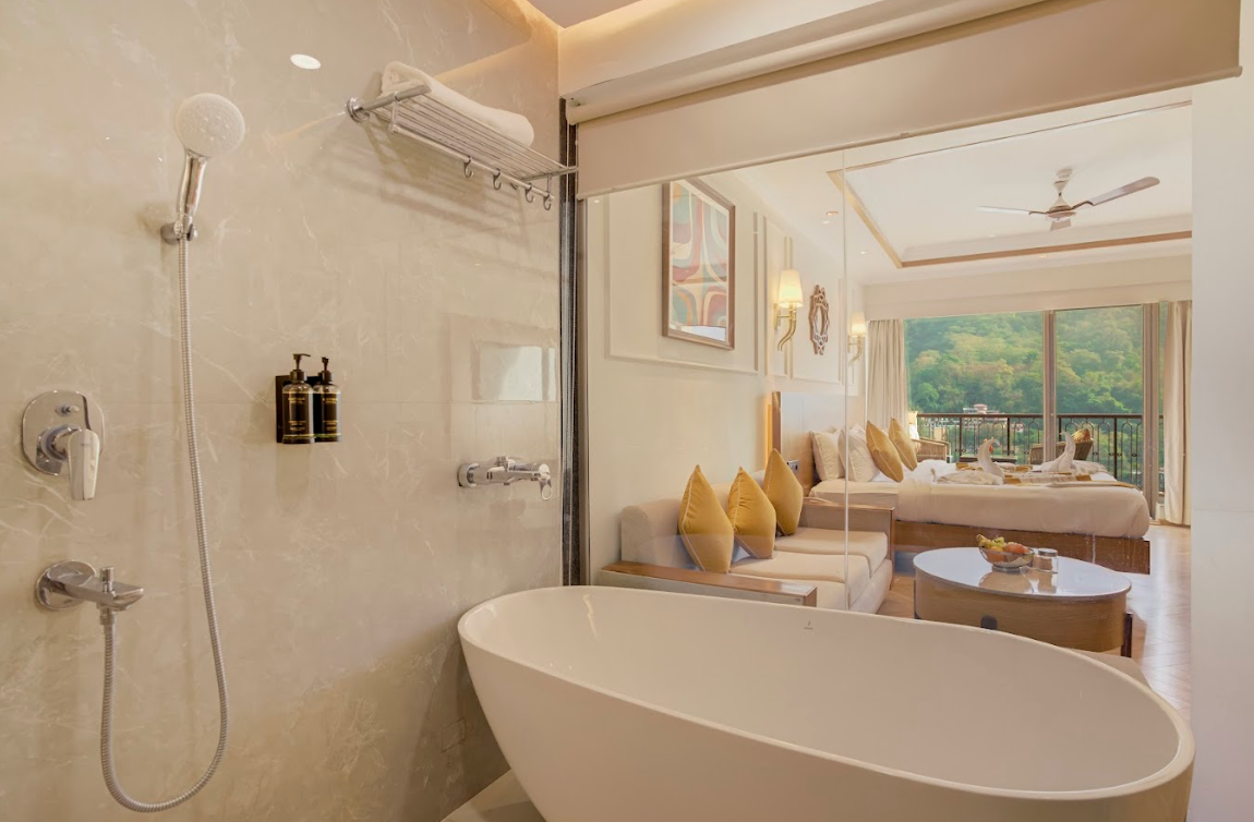 Bathroom with a white free-standing bathtub, showerhead, and toiletries, looking into a bedroom with a large bed, decorative pillows, wall art, and outdoor balcony with greenery.
