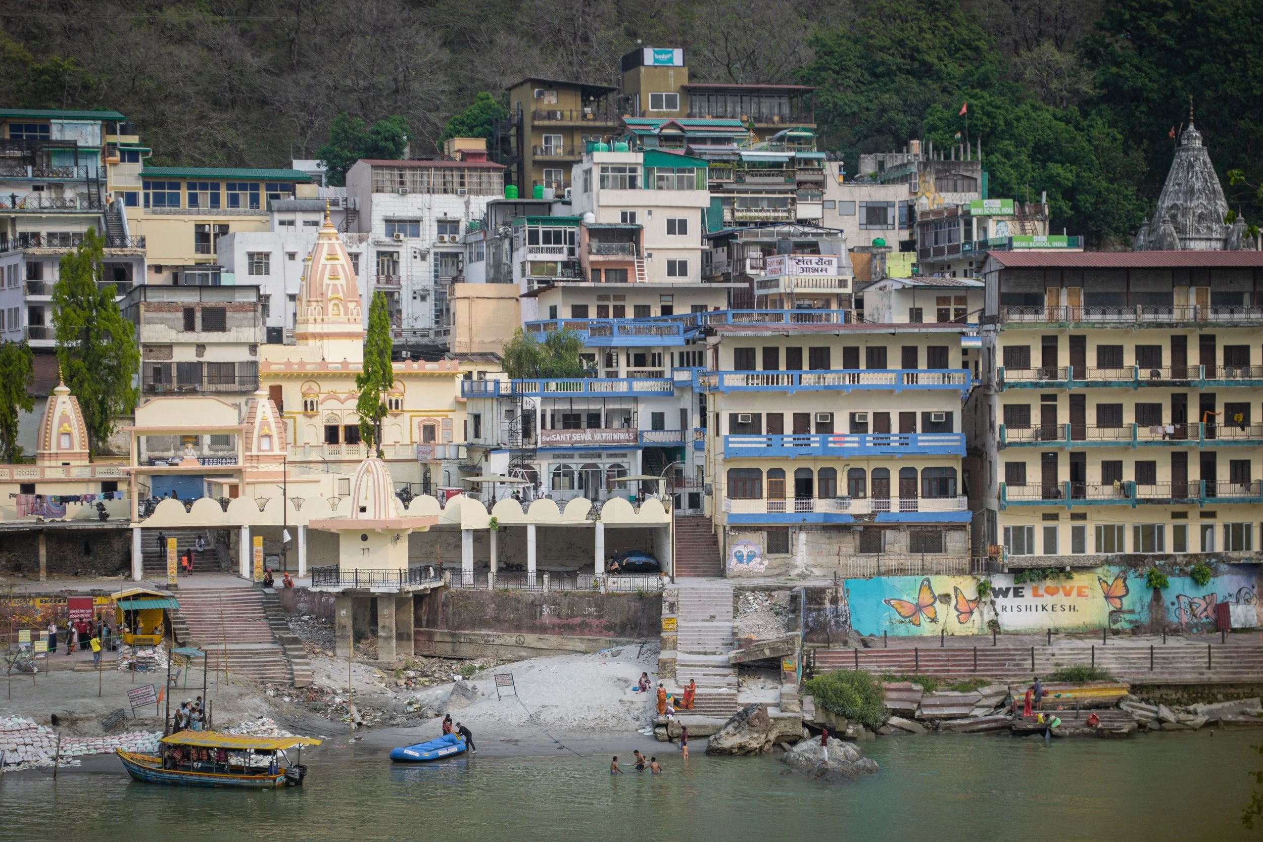 Urban hillside with multistory buildings, some with balconies, and temples with domed roofs along a riverbank, with boats and people near the water.