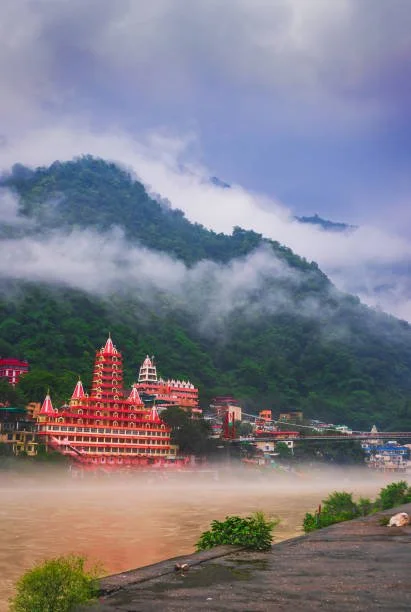 A colorful temple complex with red, pink, and white structures at the base of green, mist-covered mountains, with a river in the foreground and cloudy sky overhead.