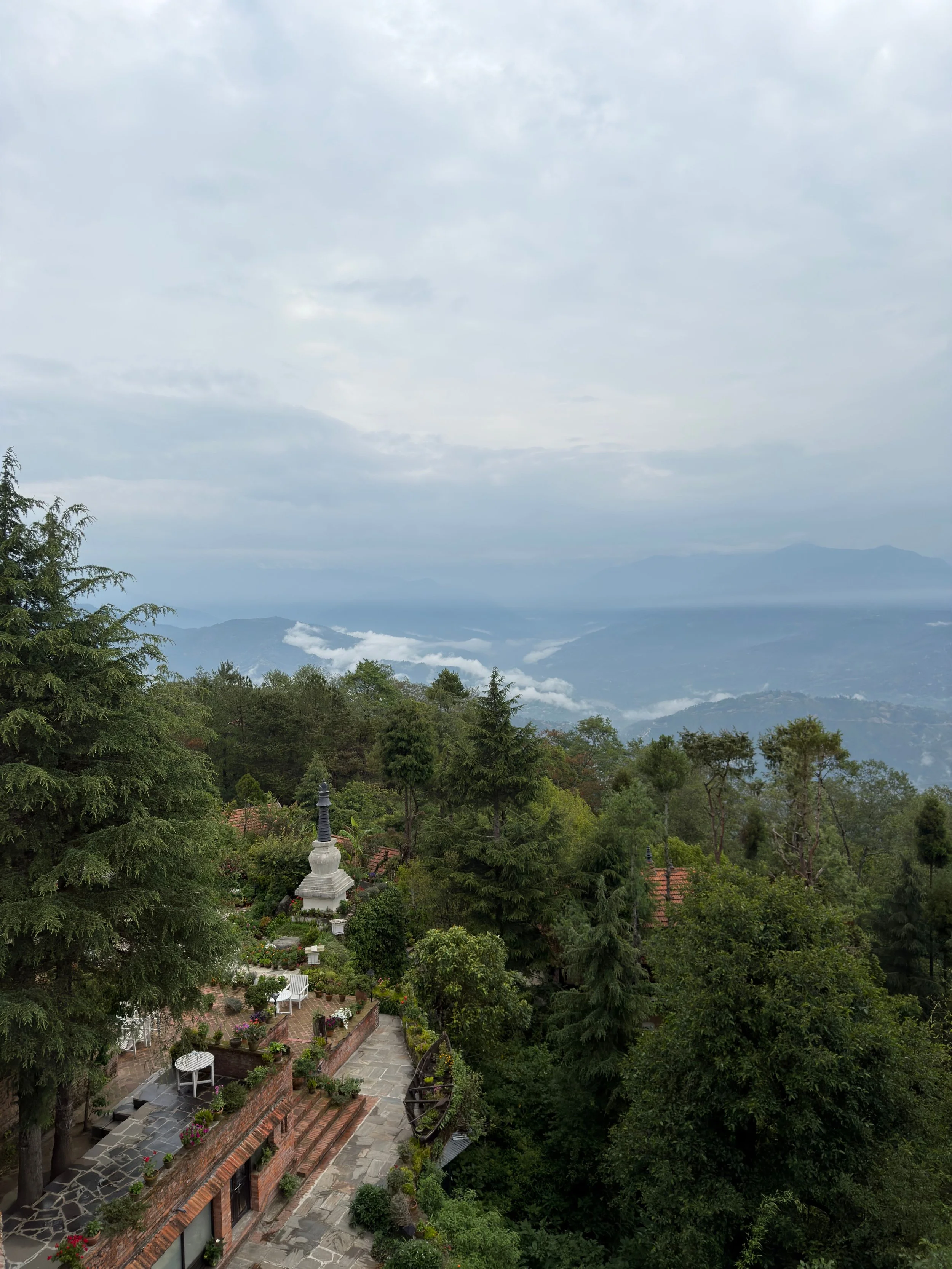 A scenic view of a hilly landscape with lush green trees, traditional buildings, and a white stupa in a garden, with mountains and cloudy sky in the background.