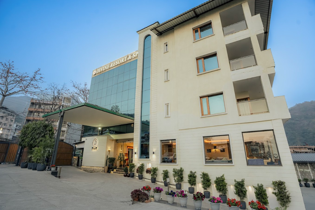 Exterior of a modern hotel building with potted plants and flowers outside, sign reads 'Divine Resort & Spa,' with mountain in the background and a clear sky.
