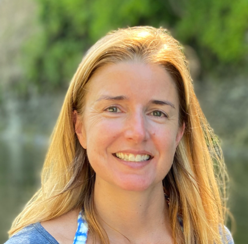 A woman with long blonde hair smiling outdoors in front of greenery and water.