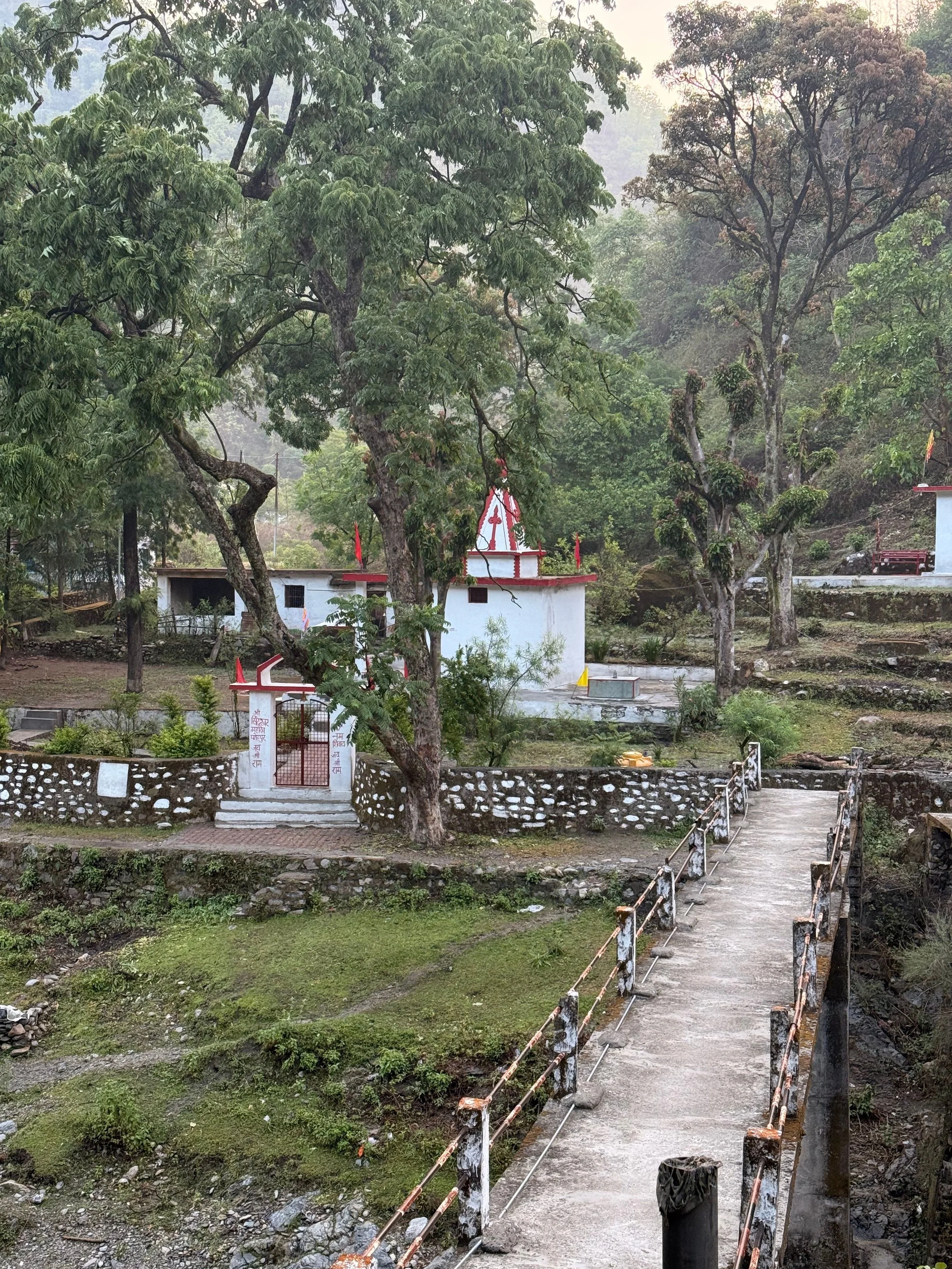 A small white temple with a red roof and decorative elements on top, situated among large trees and greenery. There is a short stairway at the entrance, surrounded by a stone fence, with a narrow footbridge leading to the temple area. The environment appears to be in a rural or forested setting.