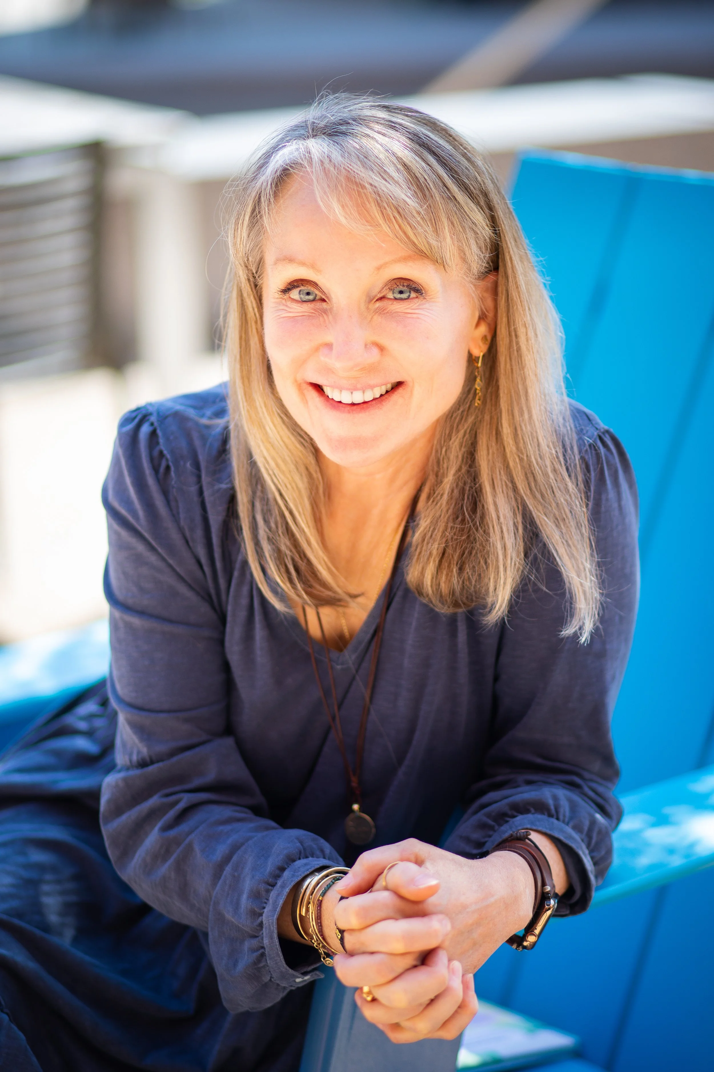 A woman with blonde hair, wearing a navy blue shirt, smiling and clasping her hands together, sitting outdoors on a blue bench.