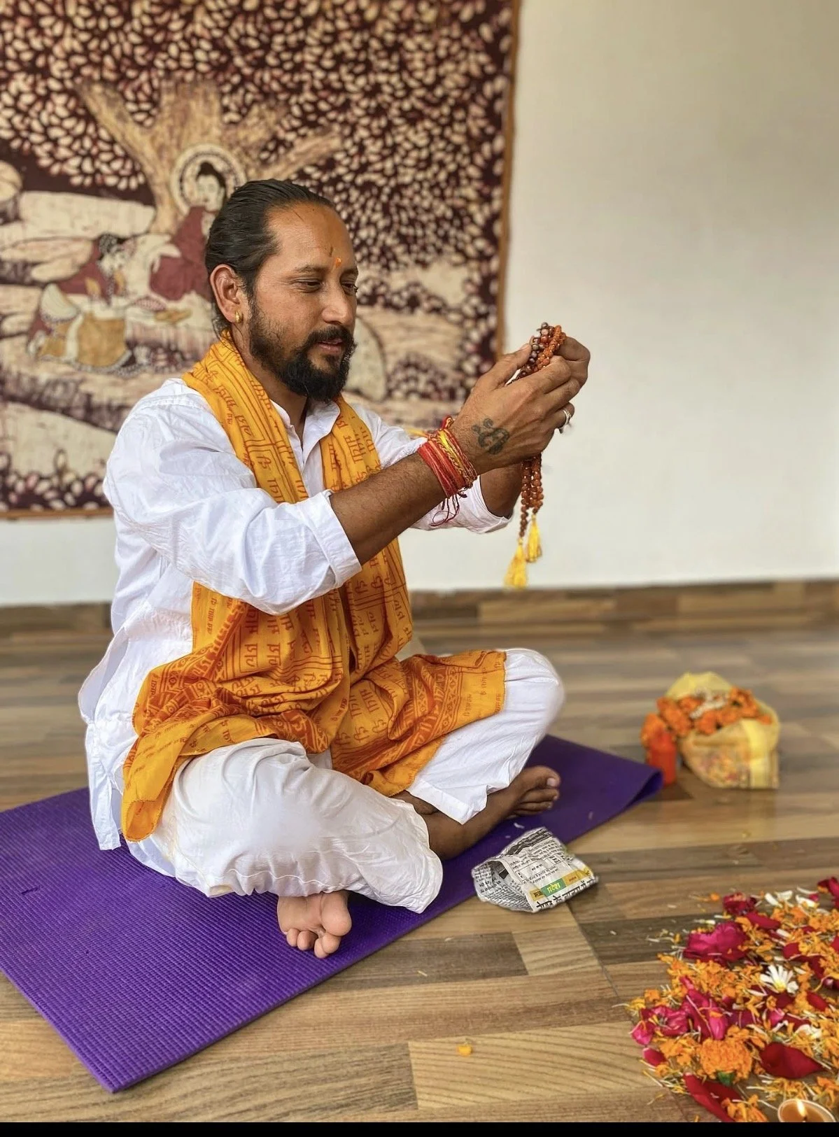 A man dressed in traditional Hindu attire, seated on a purple mat on a wooden floor, engaged in a religious ceremony with prayer beads. He has a tattoo on his hand, wears orange and white clothing with yellow religious scarves, and is surrounded by flower petals and offerings, with a tapestry featuring a deity in the background.