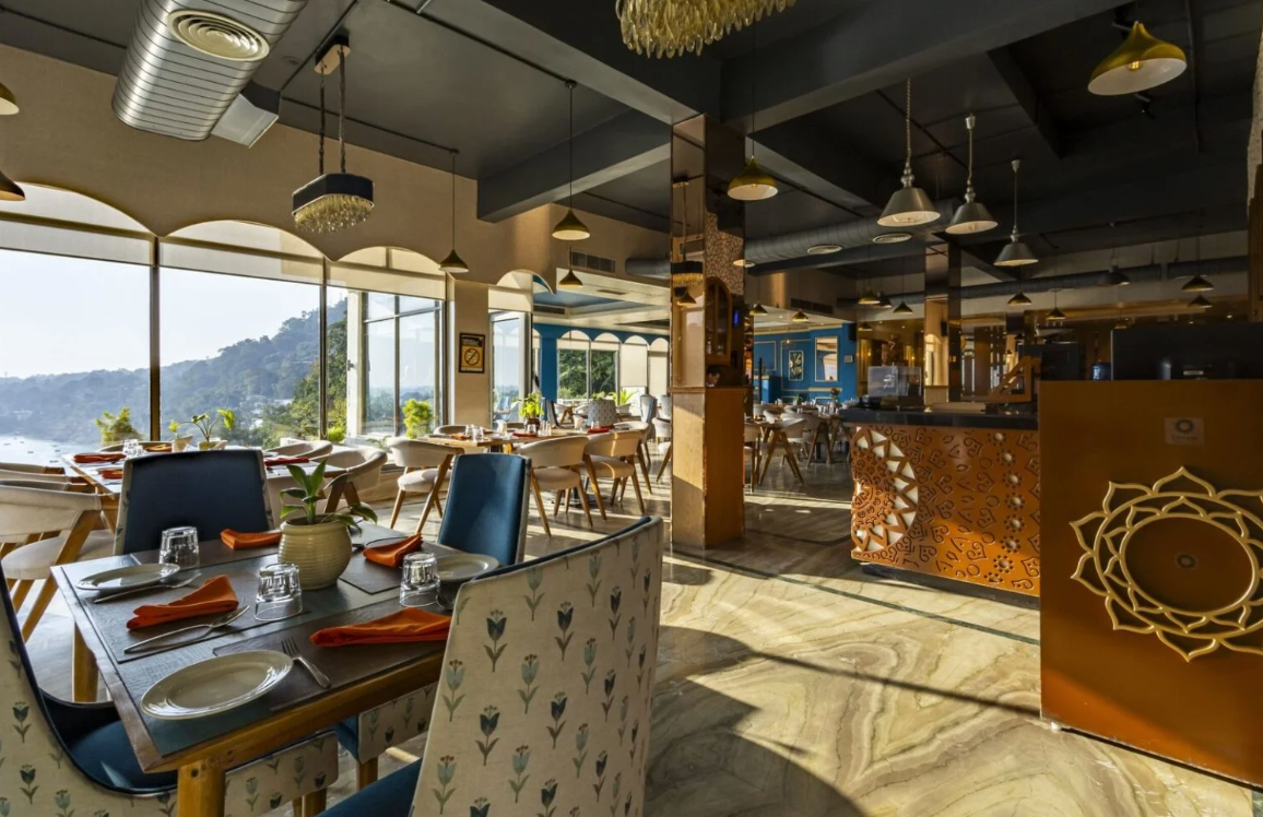 Interior of a restaurant with tables set for dining, large windows overlooking a scenic view, stylish decor, and a reception counter on the right.