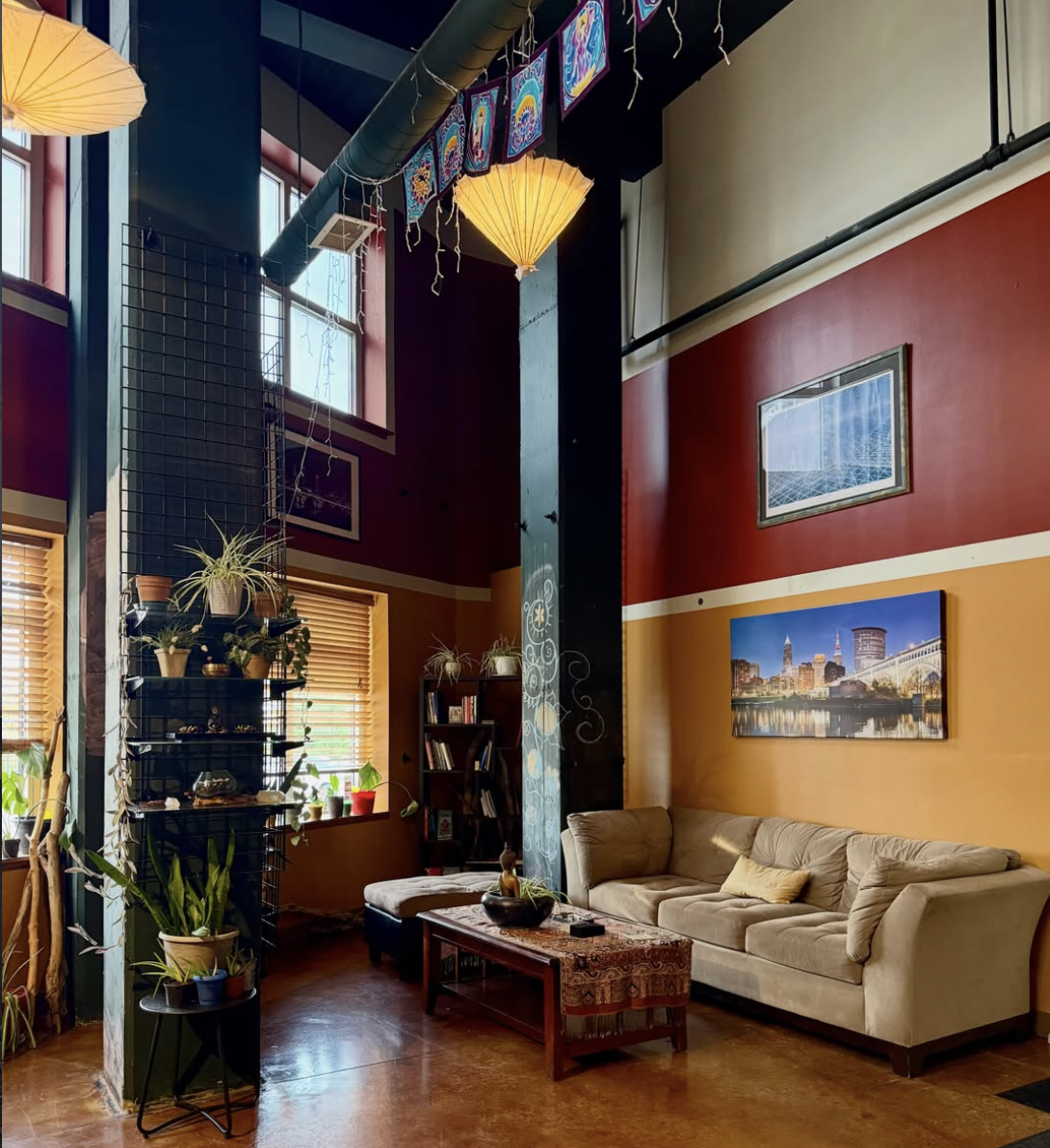A cozy living room with red and yellow walls, a beige sofa with a throw pillow, a wooden coffee table with decorative items, a bookshelf, and several potted plants. Hanging from the ceiling are colorful paper lanterns and banners with rainbow designs. Windows with wooden blinds let in natural light.