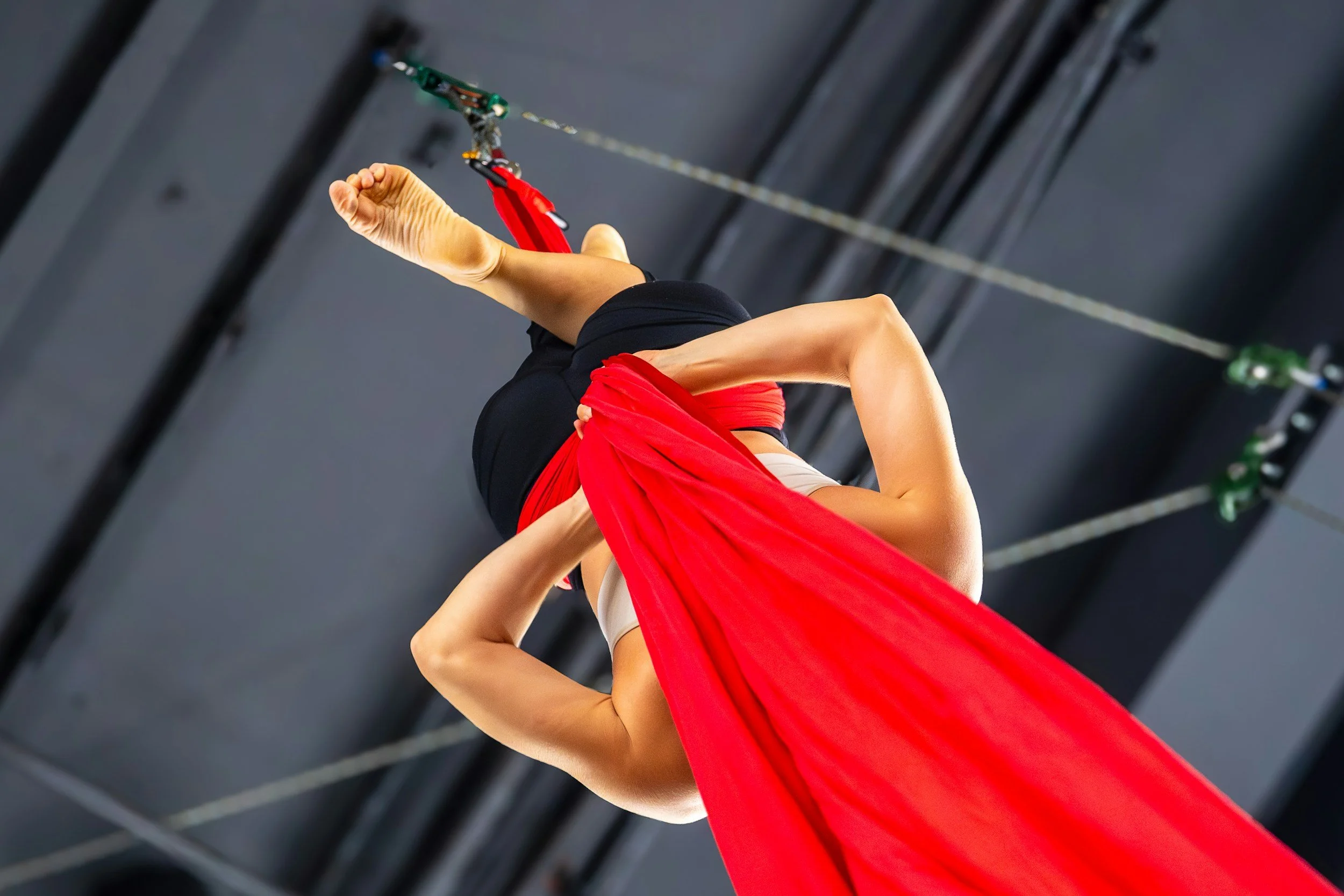 A person engaged in aerial silk acrobatics, hanging upside down with one leg wrapped around a red fabric and holding onto a harness or hardware. The setting appears to be indoors with a gray ceiling and rigging visible.