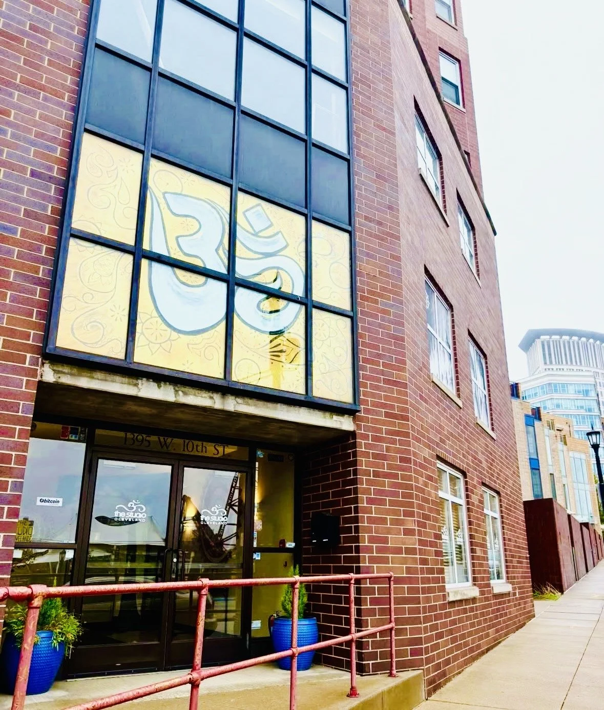 Red brick building with large windows and a yellow sign with a stylized Om symbol on the upper window, located at 1395 West 10th Street.