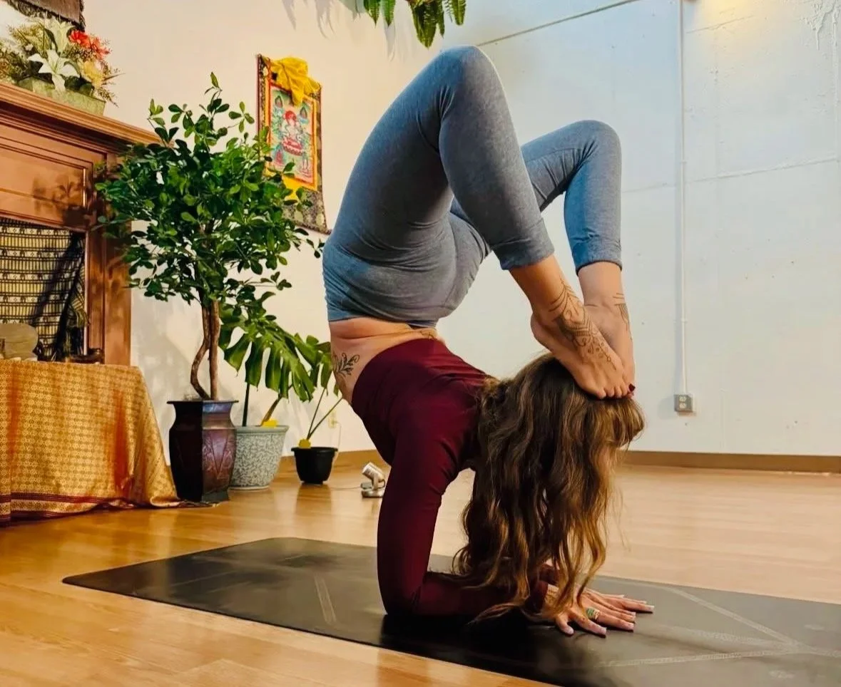 A woman practicing yoga indoors, performing a headstand with her legs bent and hands clasped behind her head, on a black yoga mat.