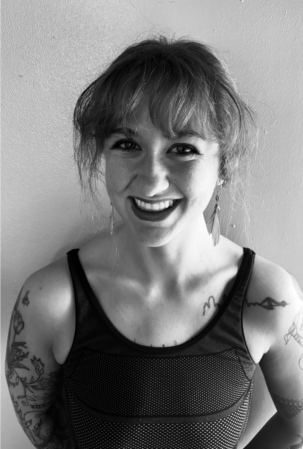 Black and white photo of a smiling woman with tattoos on her arms and shoulder, wearing a sleeveless top and earrings.