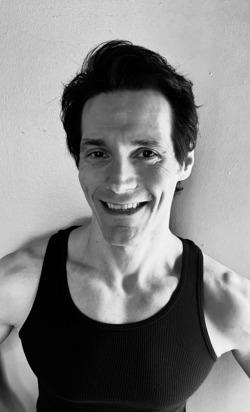 Black and white photo of a smiling man with dark, styled hair wearing a sleeveless athletic shirt, standing against a plain wall.