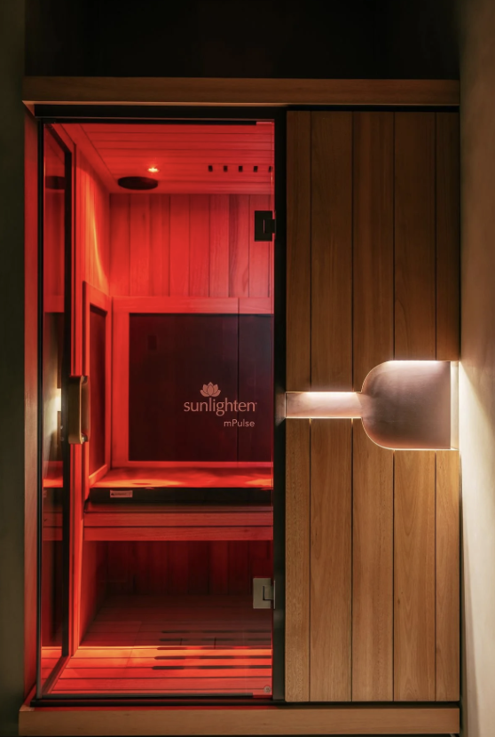 A wooden and glass sauna room with red lighting inside and a wall mounted light fixture outside.