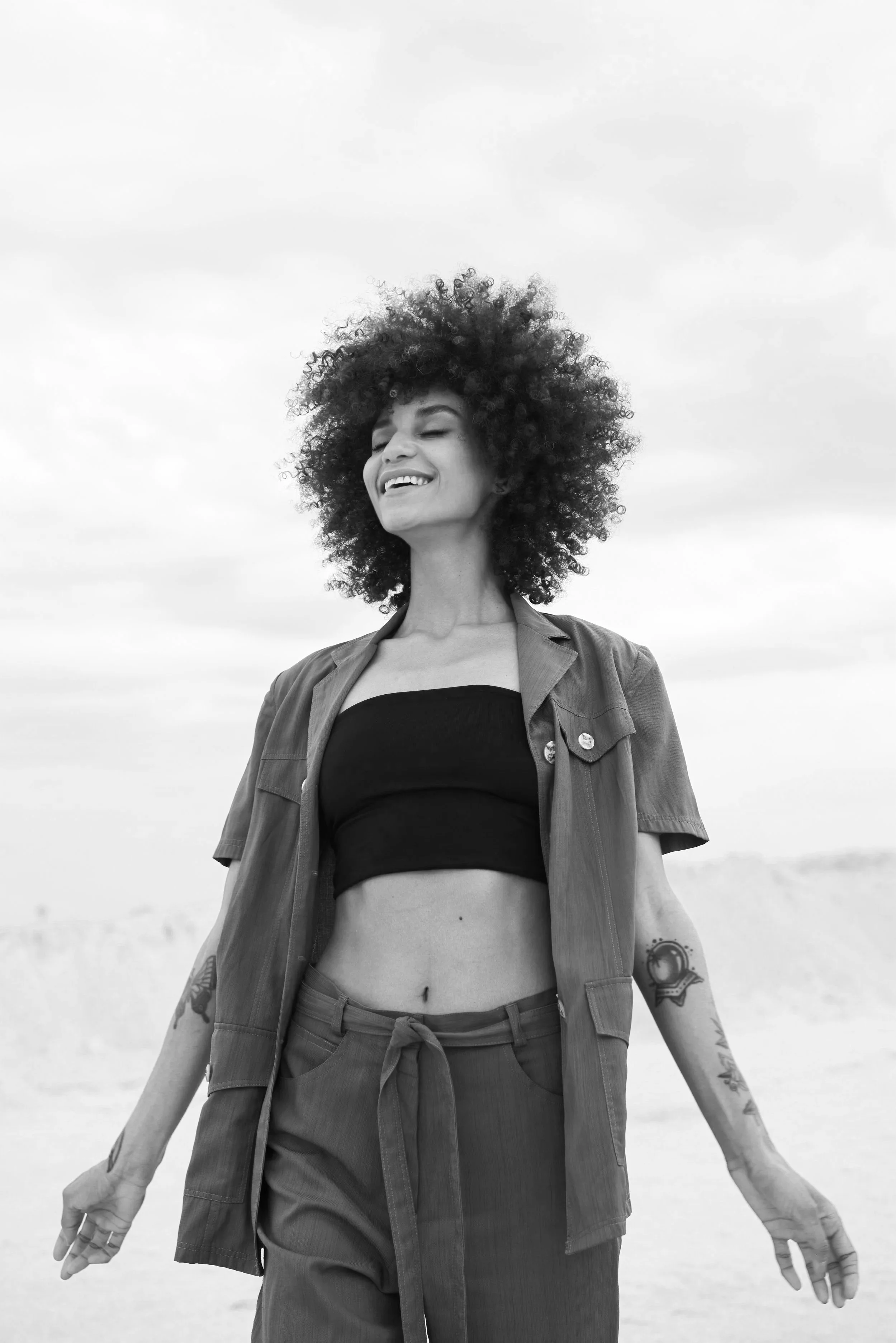 A woman with curly hair standing outdoors under a cloudy sky, smiling with eyes closed, wearing a black crop top, a casual button-up shirt, and high-waisted pants. She has tattoos on her arms.
