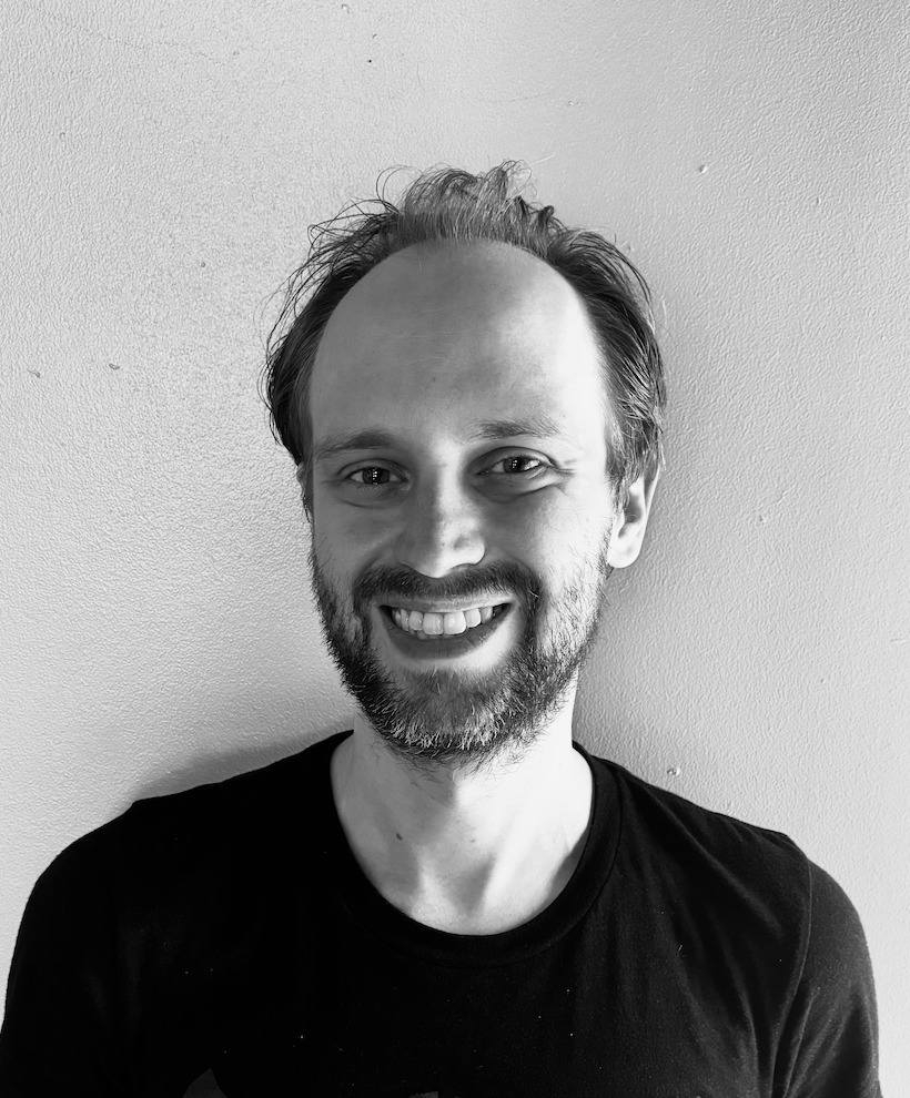 A black-and-white portrait of a smiling man with short, slightly messy hair, a beard, and wearing a black t-shirt, standing against a plain wall.