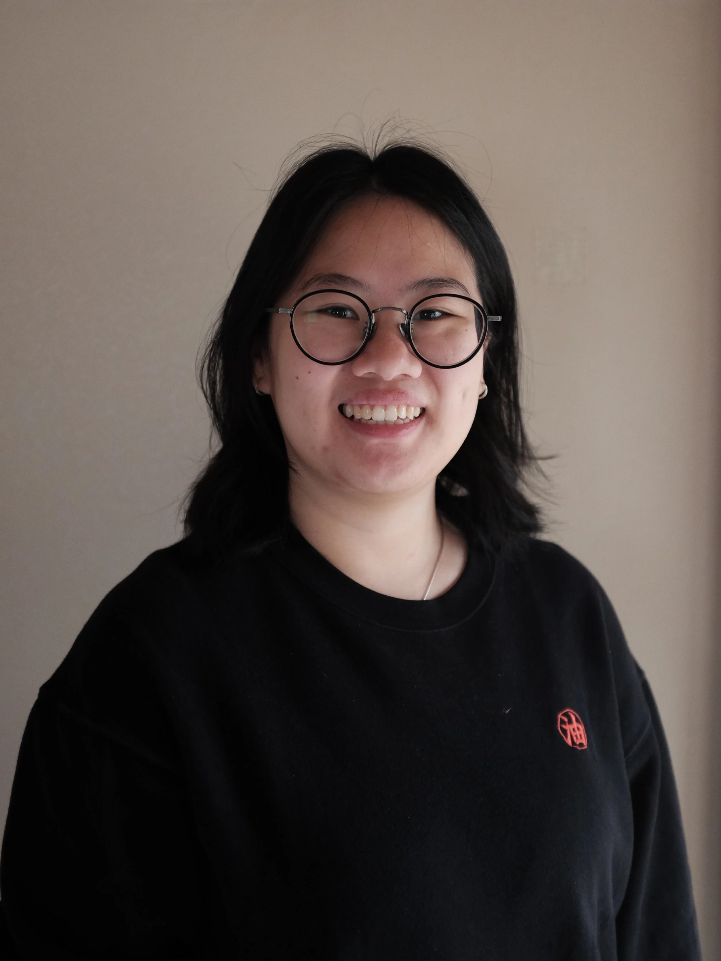 A young woman with short black hair, wearing glasses and a black sweatshirt with a red logo, smiling in front of a plain wall.