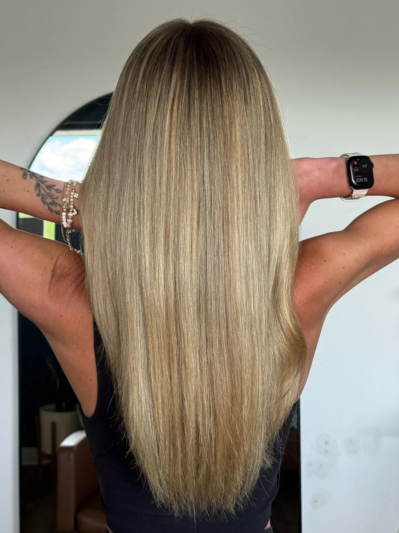 A woman with long, straight, blonde hair taking a mirror selfie, wearing a smartwatch and a tattooed arm with jewelry.