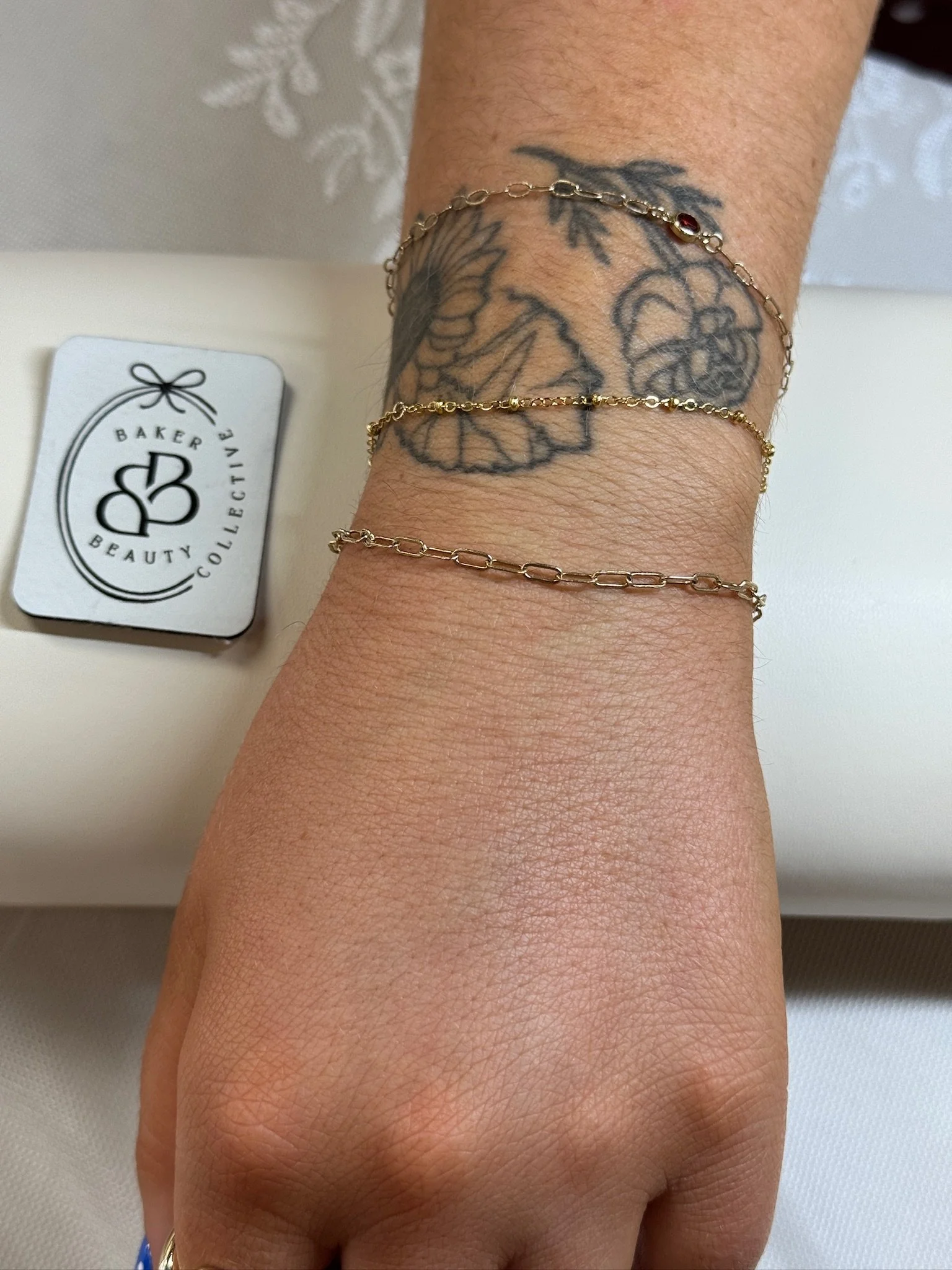 A person's arm with a tattoo of a skull and floral design, wearing three gold chain bracelets, with a Baked Beauty Collective card nearby.