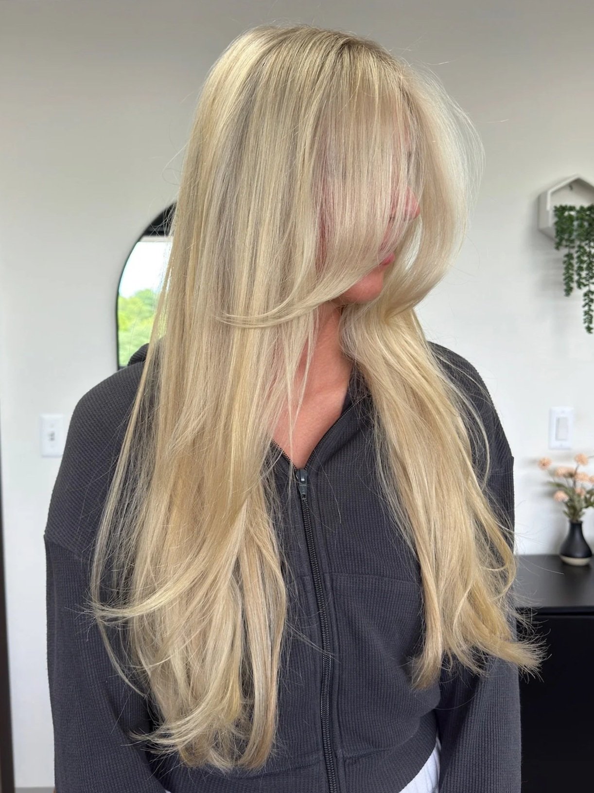 Woman with long, blonde hair styled in soft waves, wearing a black zip-up jacket, in a modern room with minimal decor.