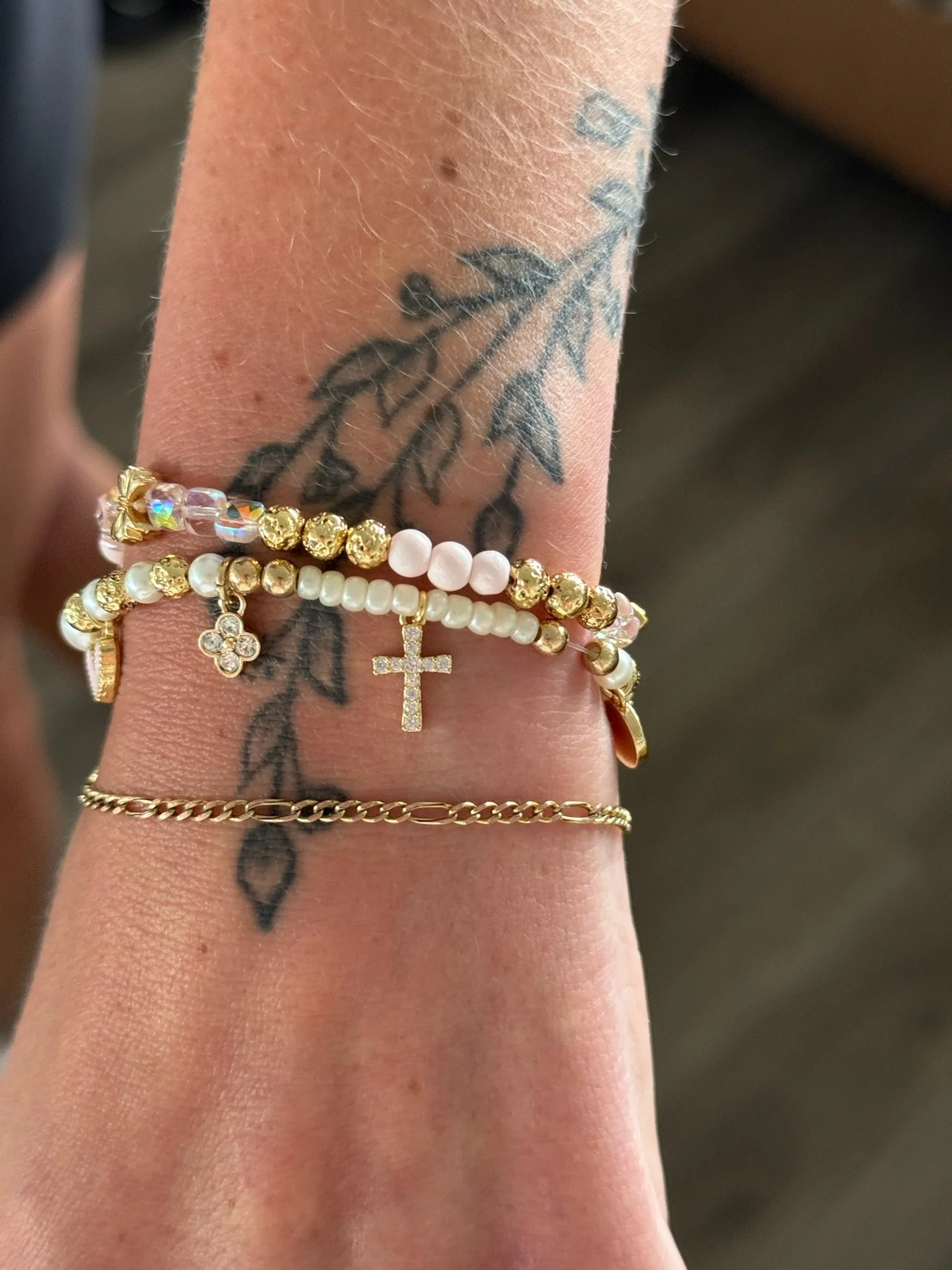 A person's forearm with several tattoos and wearing multiple bracelets, including a gold chain bracelet, a beaded bracelet with a small cross charm, and another bracelet with pink and gold beads.