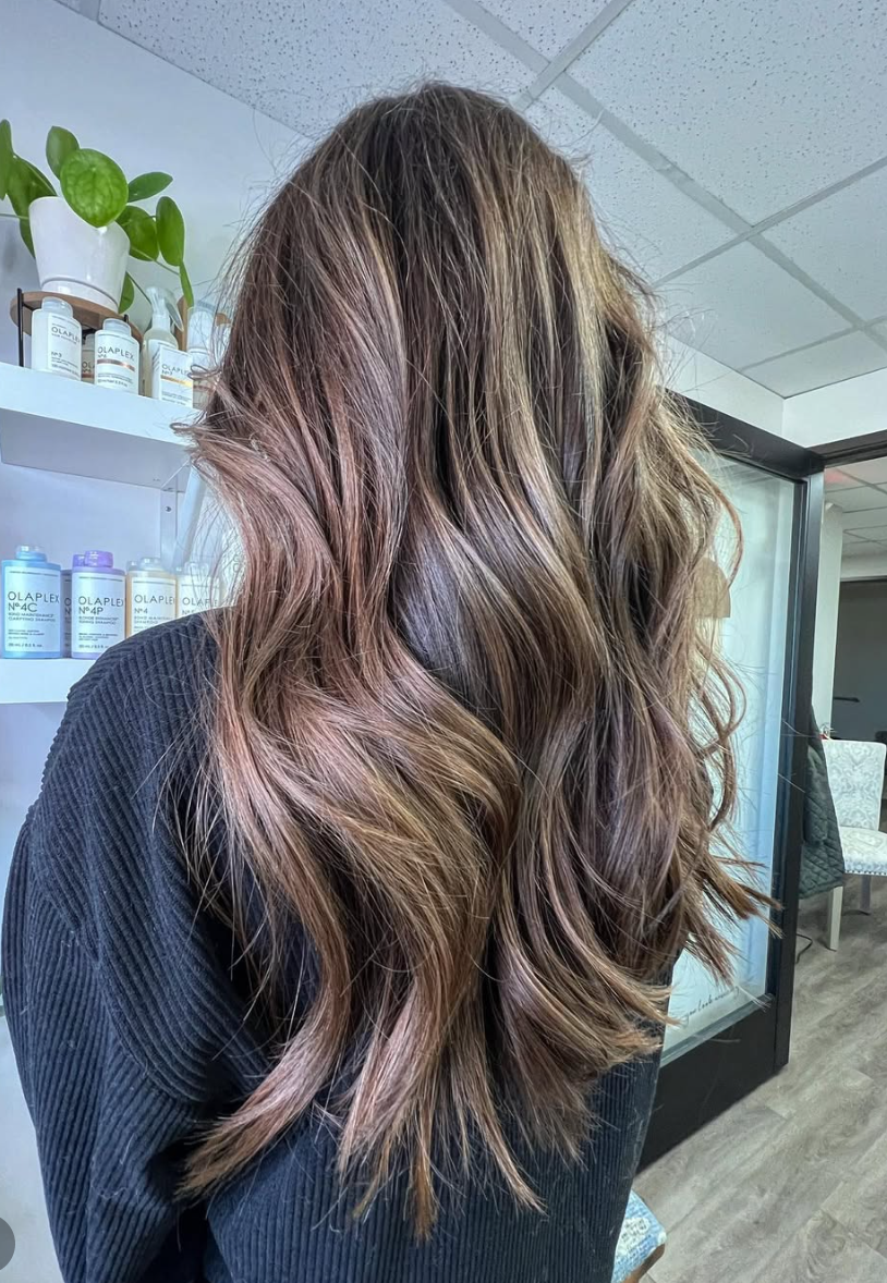 A woman with long, wavy brown hair with subtle highlights standing indoors, with shelves of hair products and plants in the background.