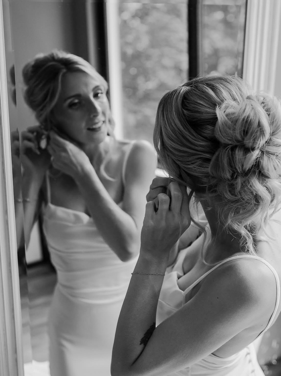 Black and white photo of two women with styled hair and dresses, one looking into a mirror and the other touching her face, in a room with a window in the background.