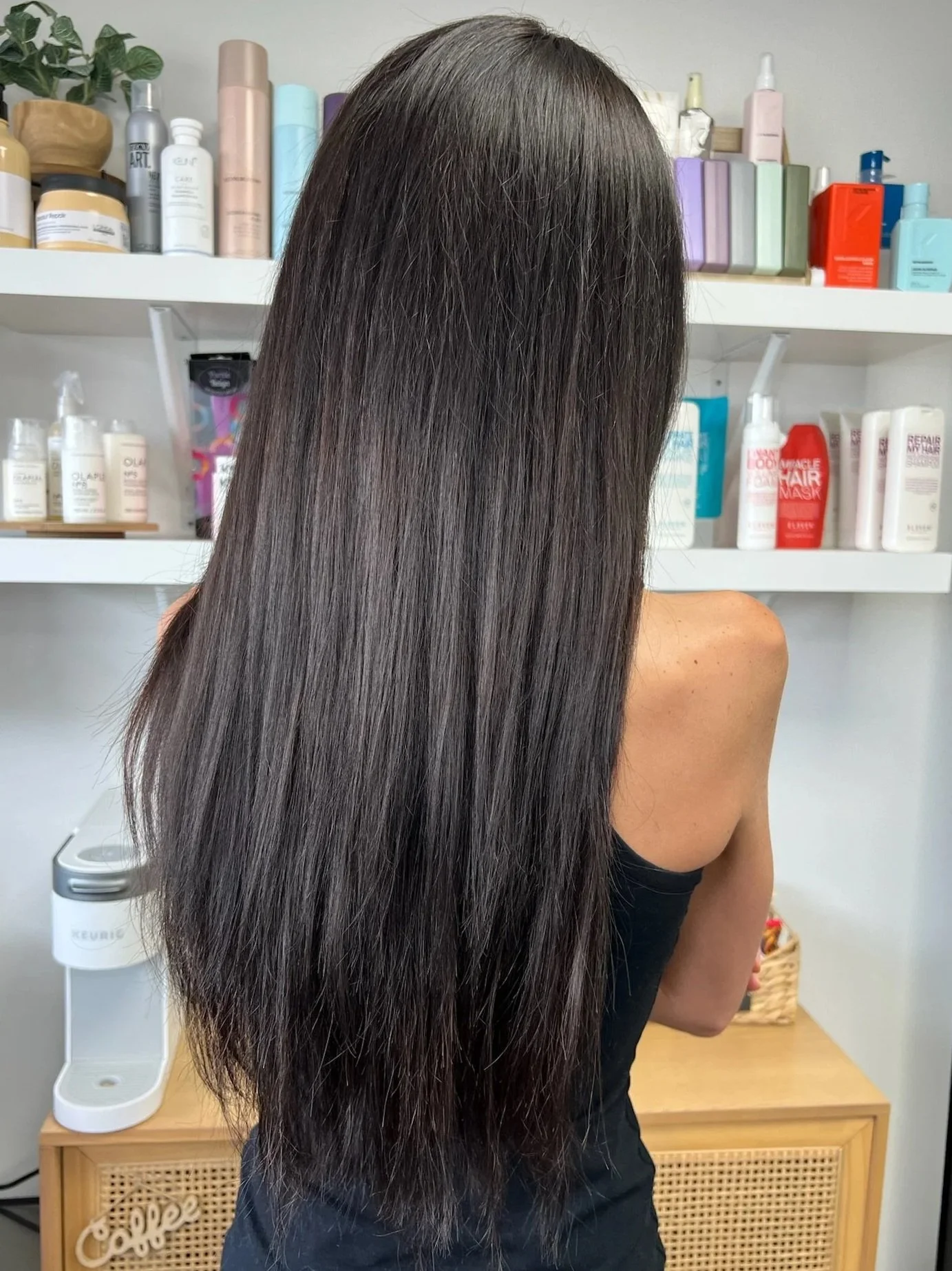 A woman with long, straight, brown hair standing in front of shelves containing hair and skincare products.