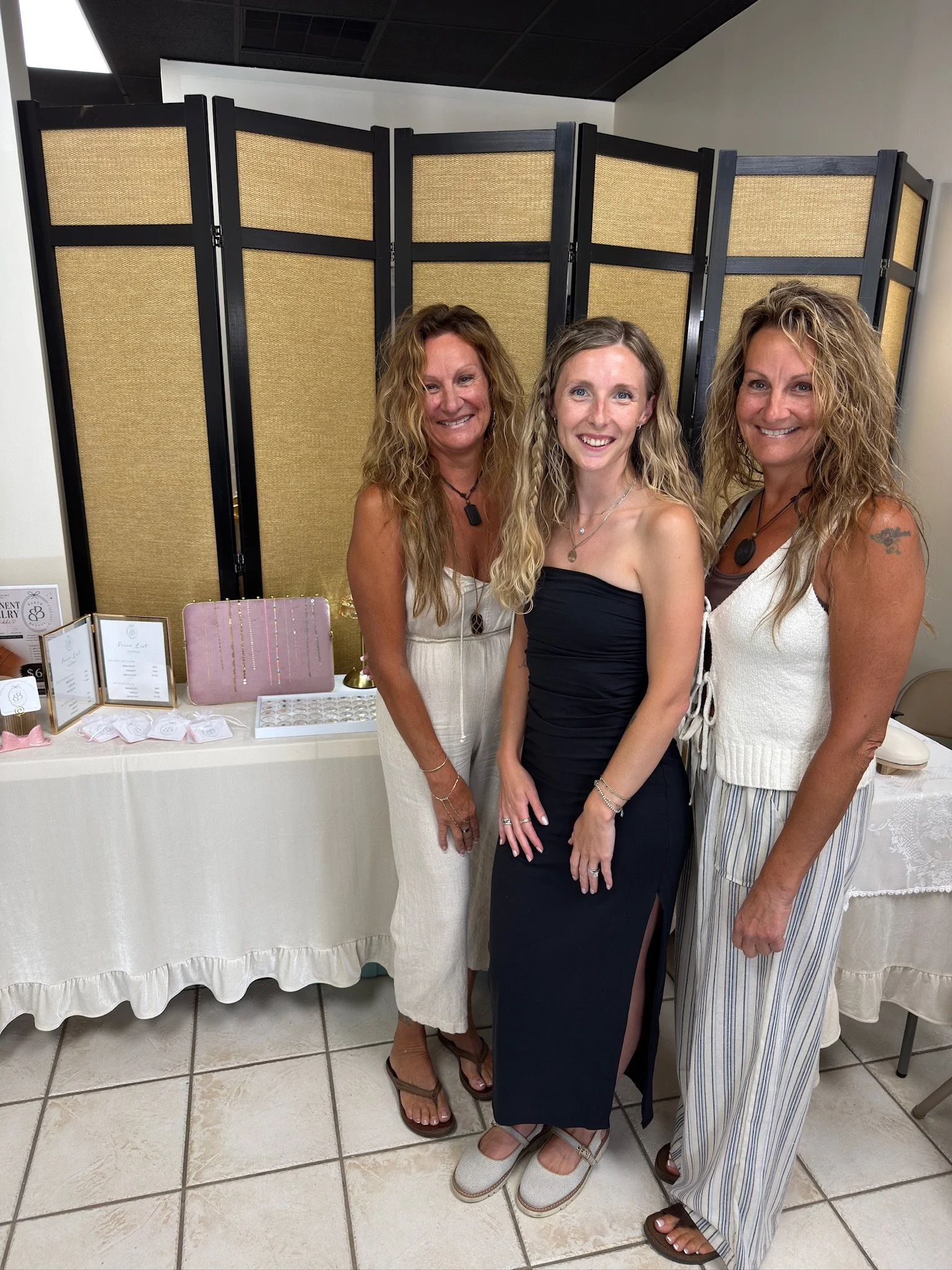 Three women standing close together at an indoor event, posing for the photo with tables displaying jewelry and accessories behind them.
