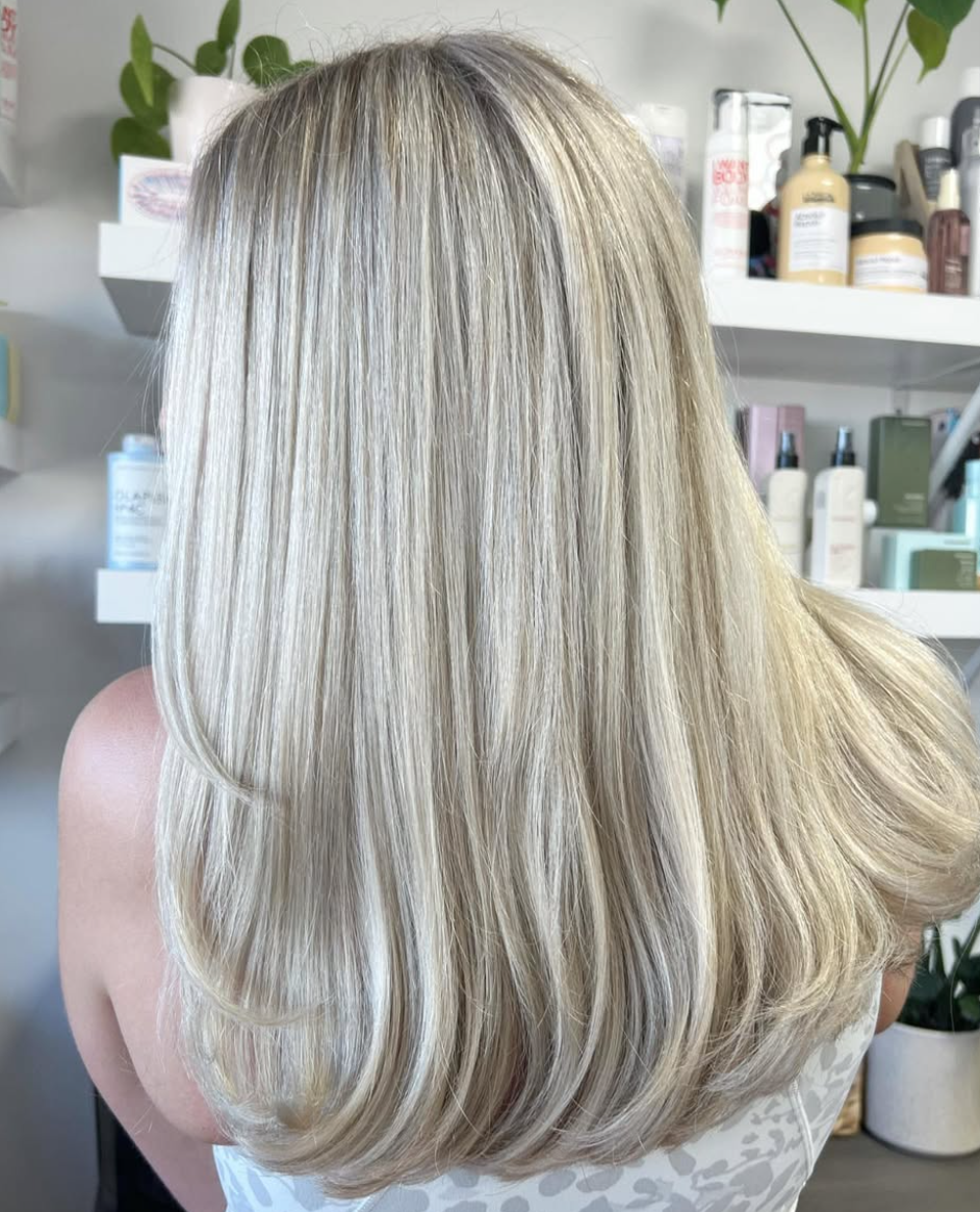 Back of a woman with long, straight, blonde hair in a salon with various hair care products on shelves.