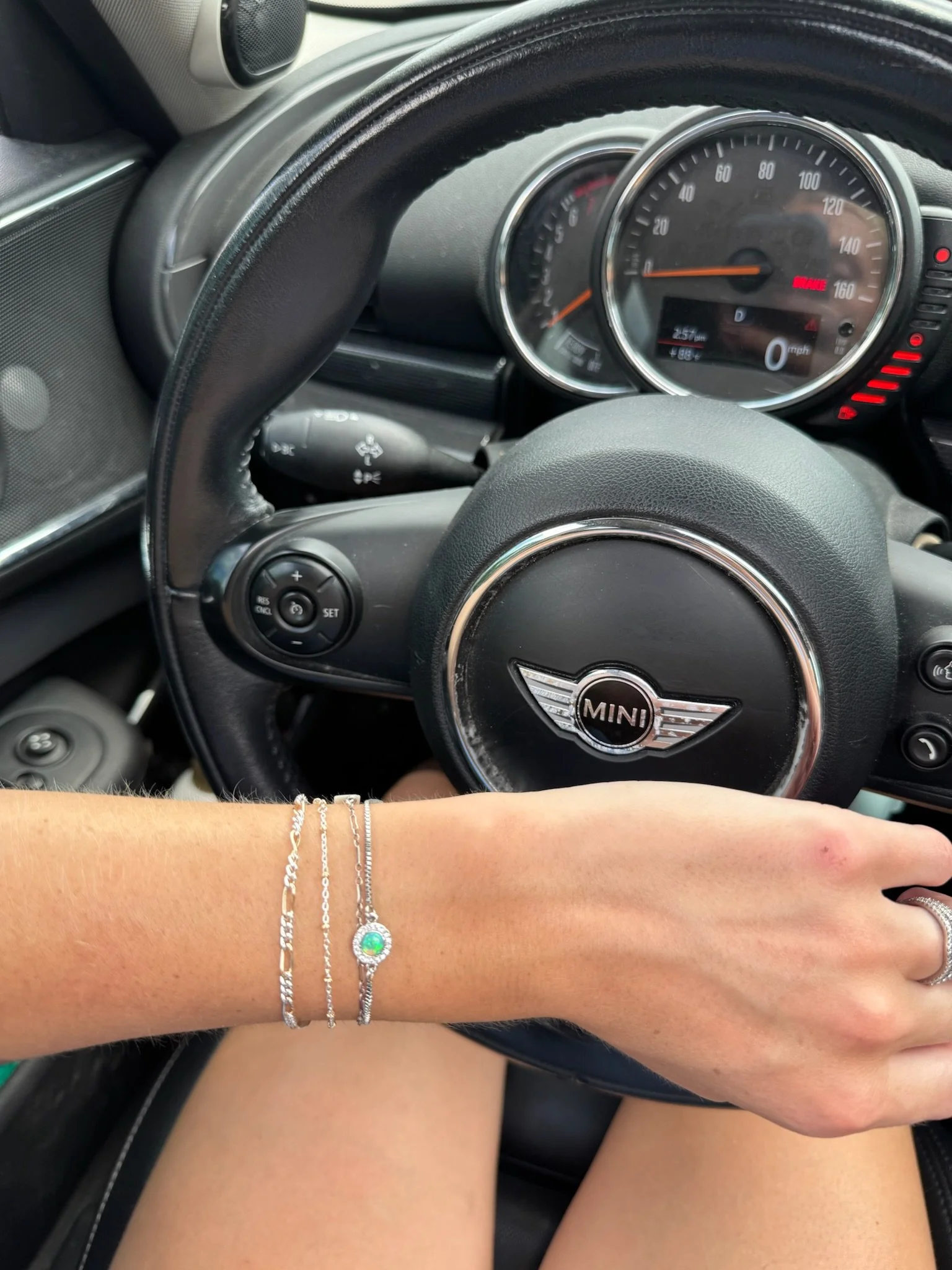 A person's arm with three silver bracelets resting on a black Mini Cooper steering wheel, with the speedometer and dashboard visible in the background.