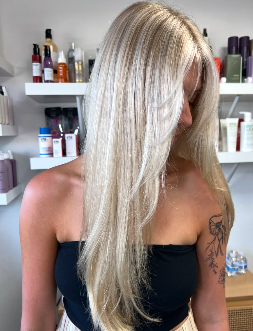 A woman with long, platinum blonde hair in a salon. She wears a black strapless top, and a tattoo of flowers is visible on her left shoulder. Behind her are shelves with hair and skincare products.